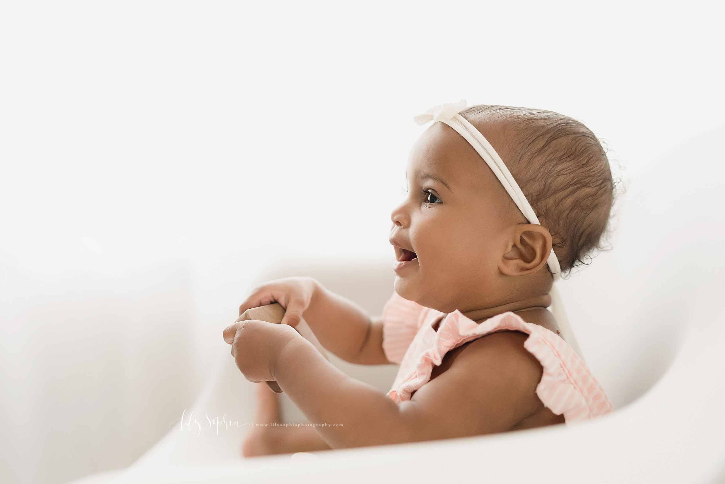  Profile milestone photo of an African-American baby girl as she sits in a white molded chair in front of a natural light window and plays with a wooden stacking toy in Atlanta. 