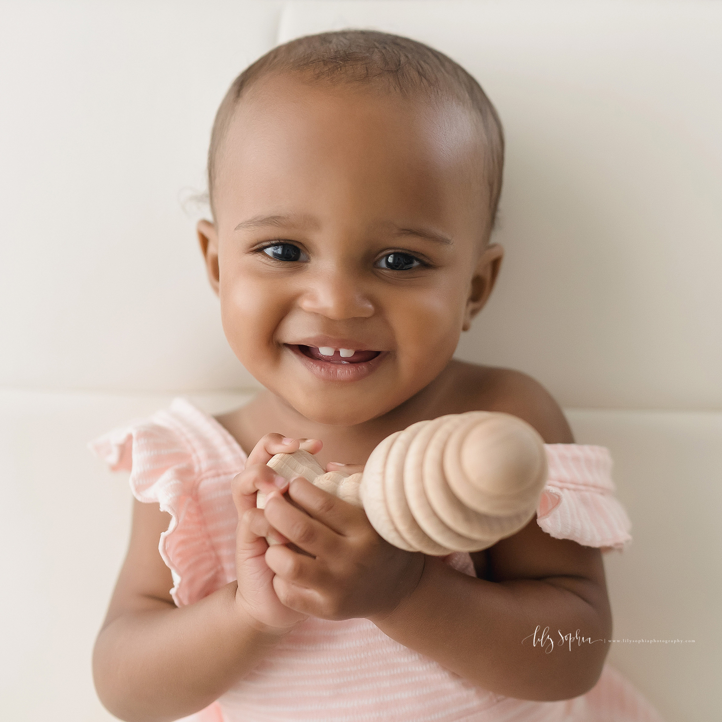  Milestone photo of a happy African-American baby girl who is one as she smiles with her teeth showing and holds a wooden toy in natural light in an Atlanta studio. 