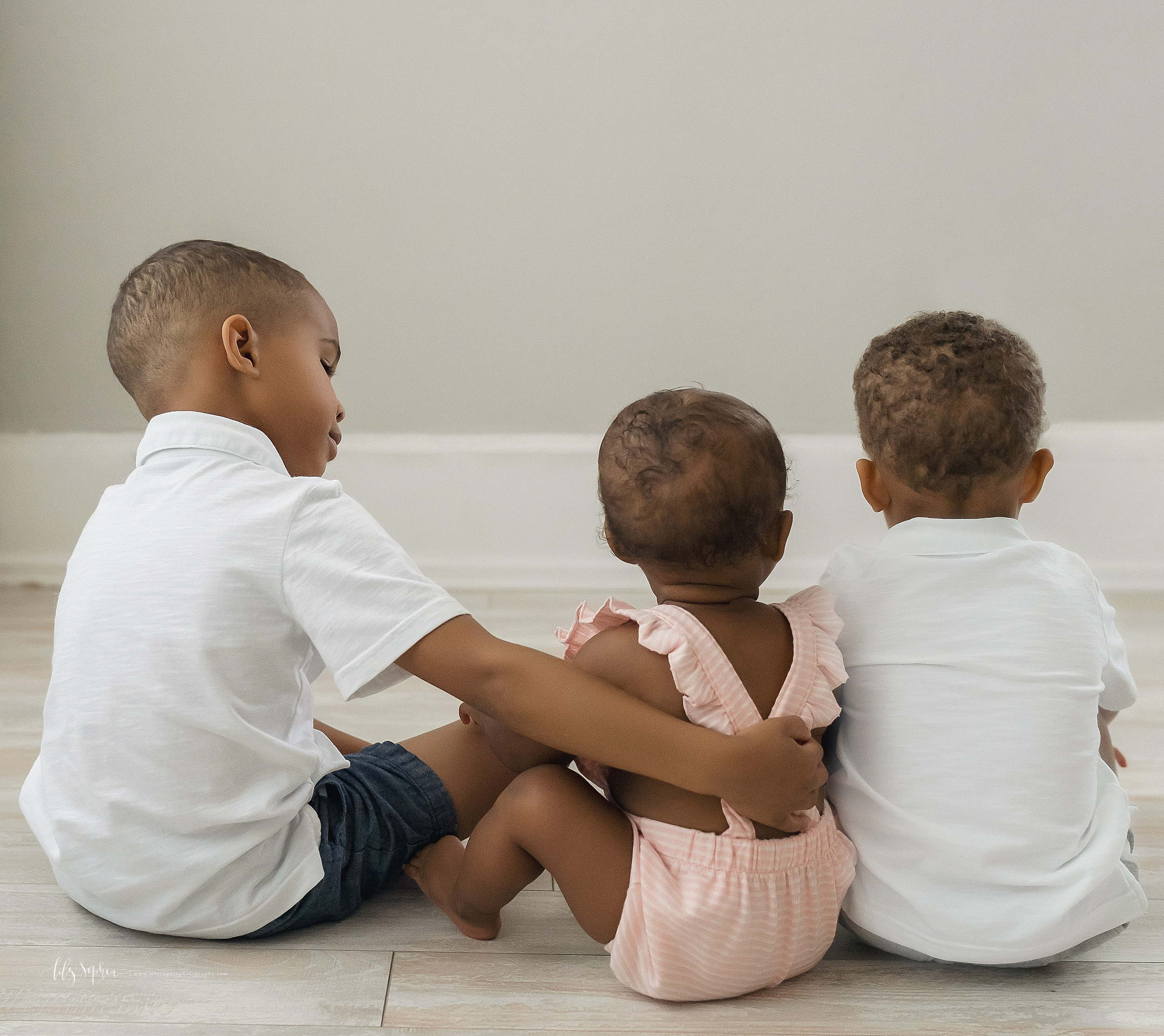  Family photo of African-American siblings as the one year old sister sits between the two brothers on the floor of an Atlanta natural light studio. 