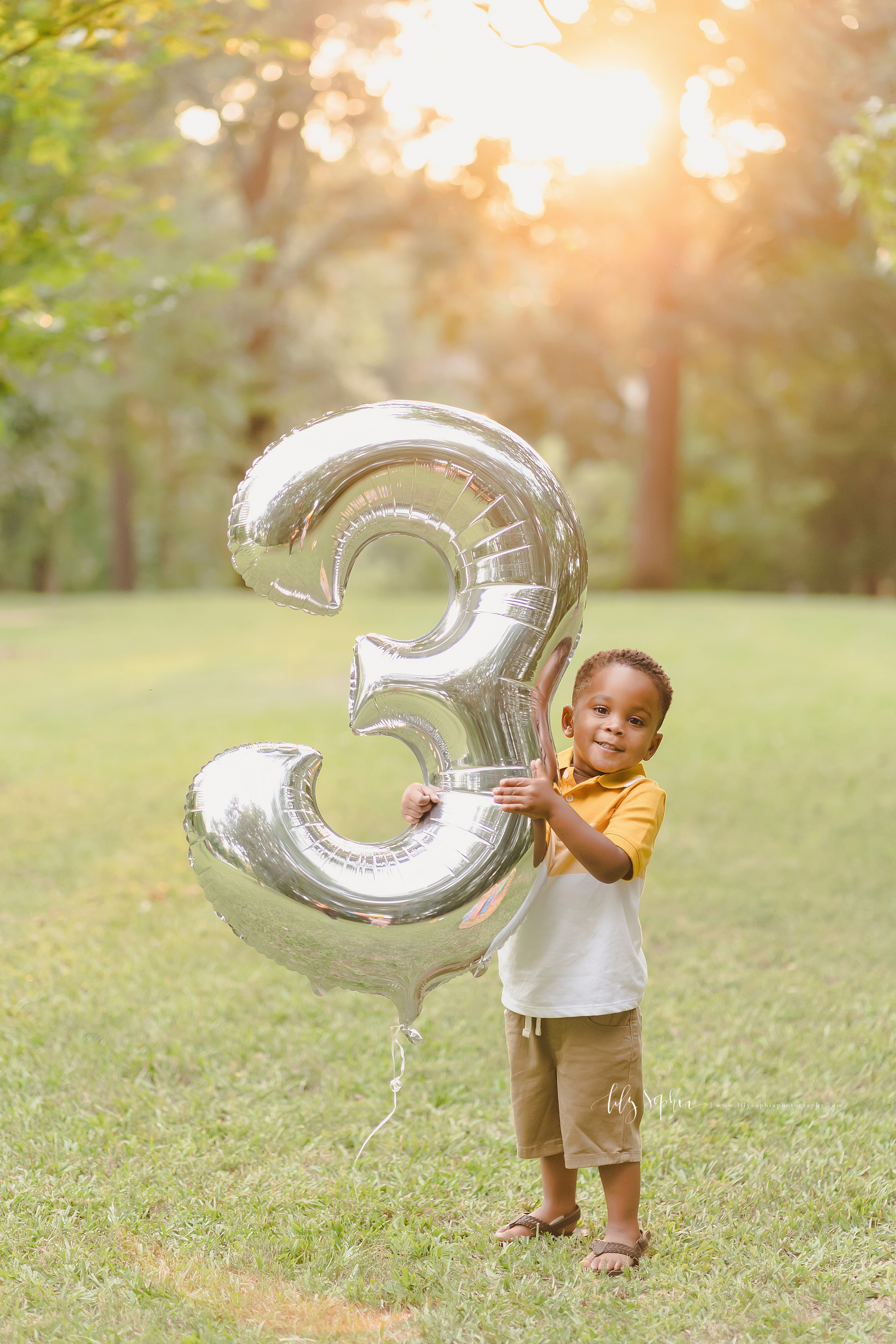  Milestone photo of an African-American boy holding a large silver number 3 balloon as he celebrates his birthday in a park at sunset in Atlanta. 