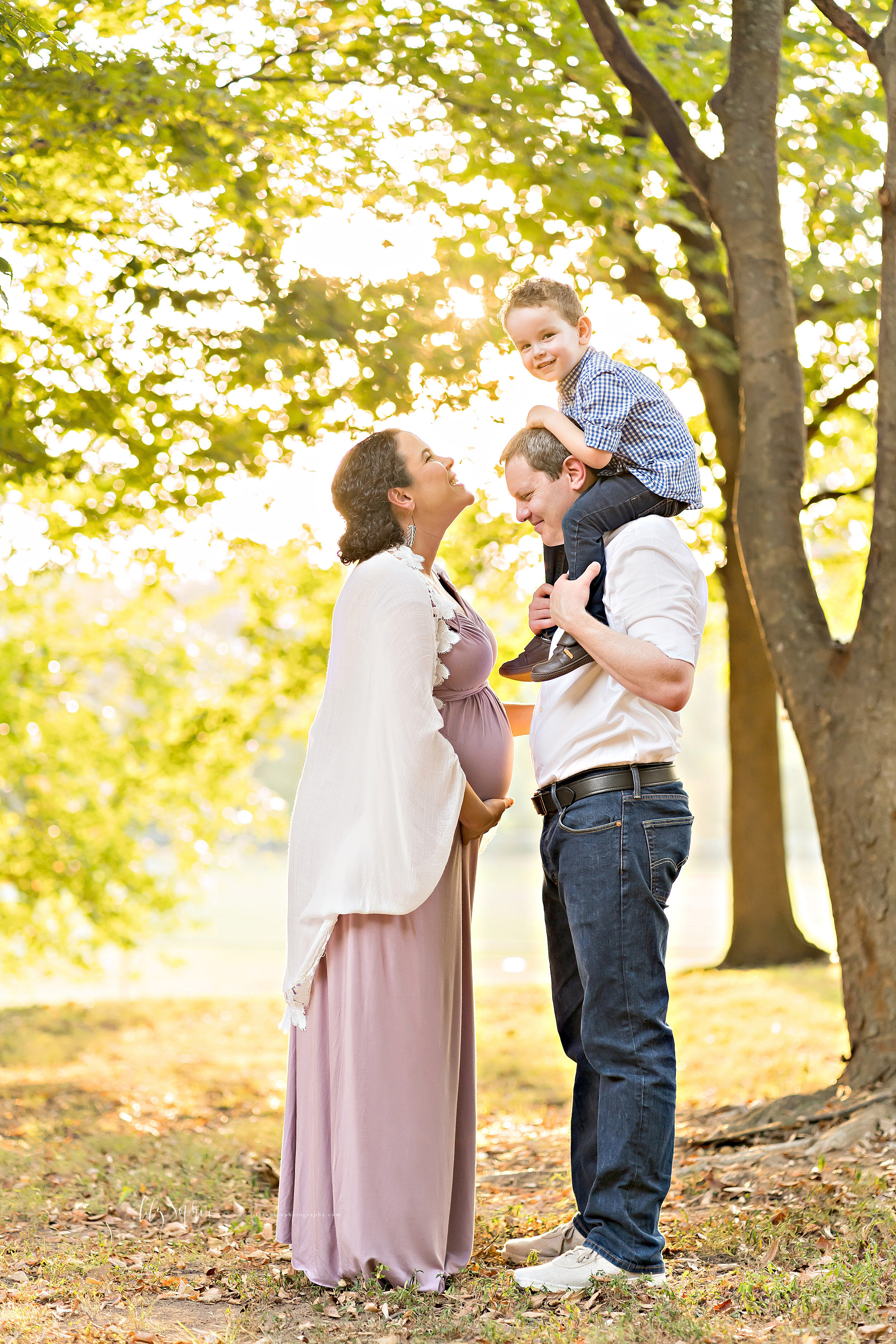  Family photo of three with expectant mom, dad, and toddler son taken in an Atlanta park at sunset as pregnant mom faces dad who has their toddler son on his shoulders. 