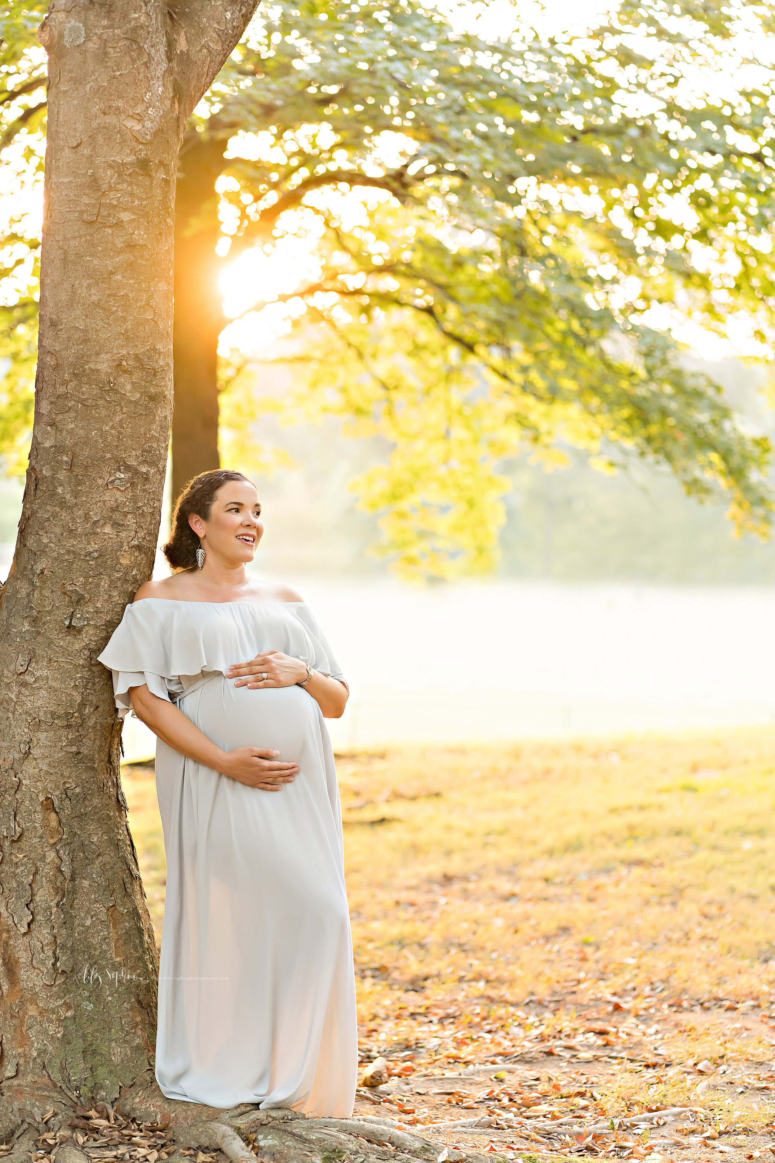  Maternity photo of a contented mother in an Atlanta park with a lake in the background who is framing her belly as she stands next to a tree at sunset. 