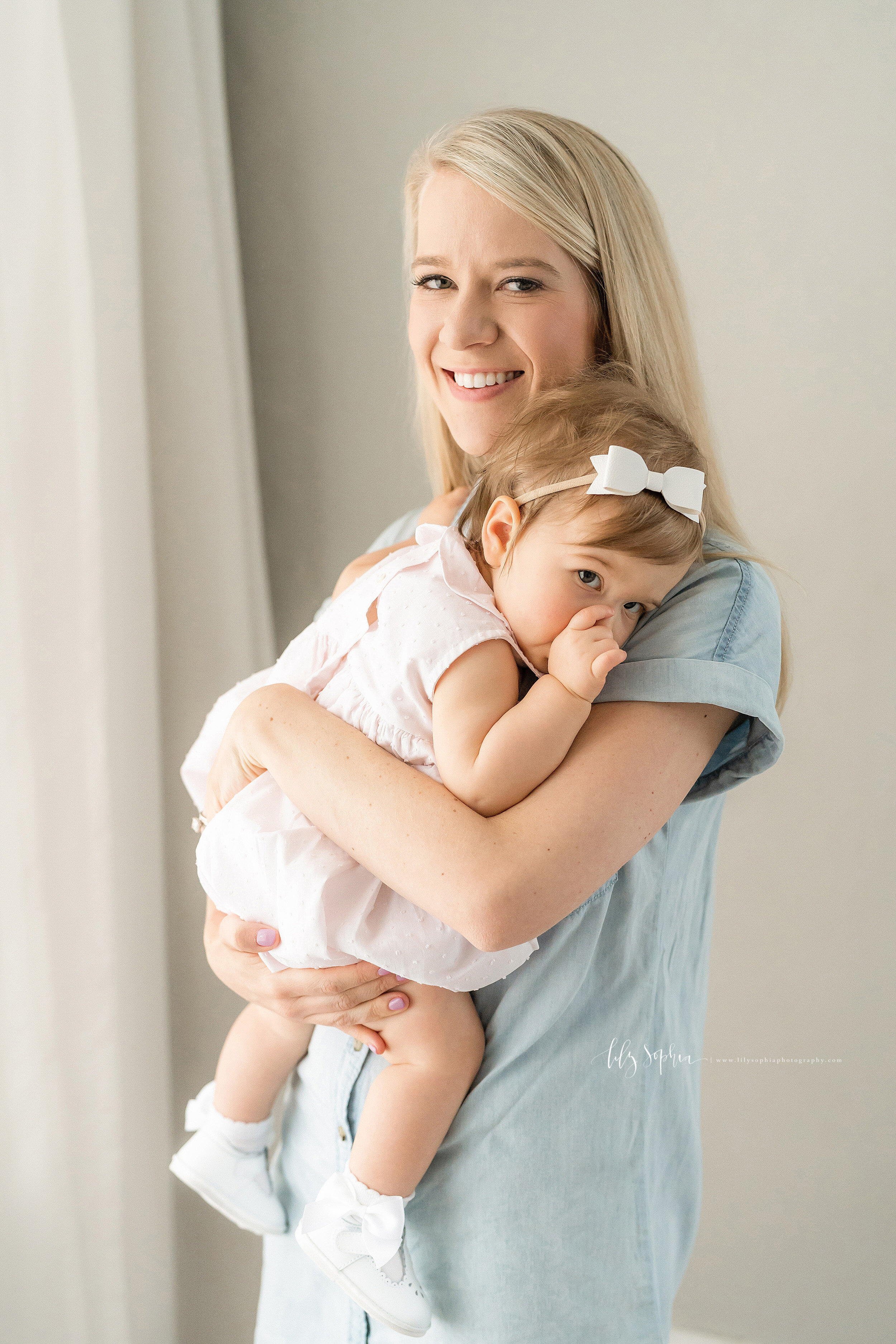  Family photo of a happy, smiling mother standing in natural light in front of a window as she holds her one year old daughter who is sucking her thumb taken in a studio in the Brookhaven area of Atlanta. 
