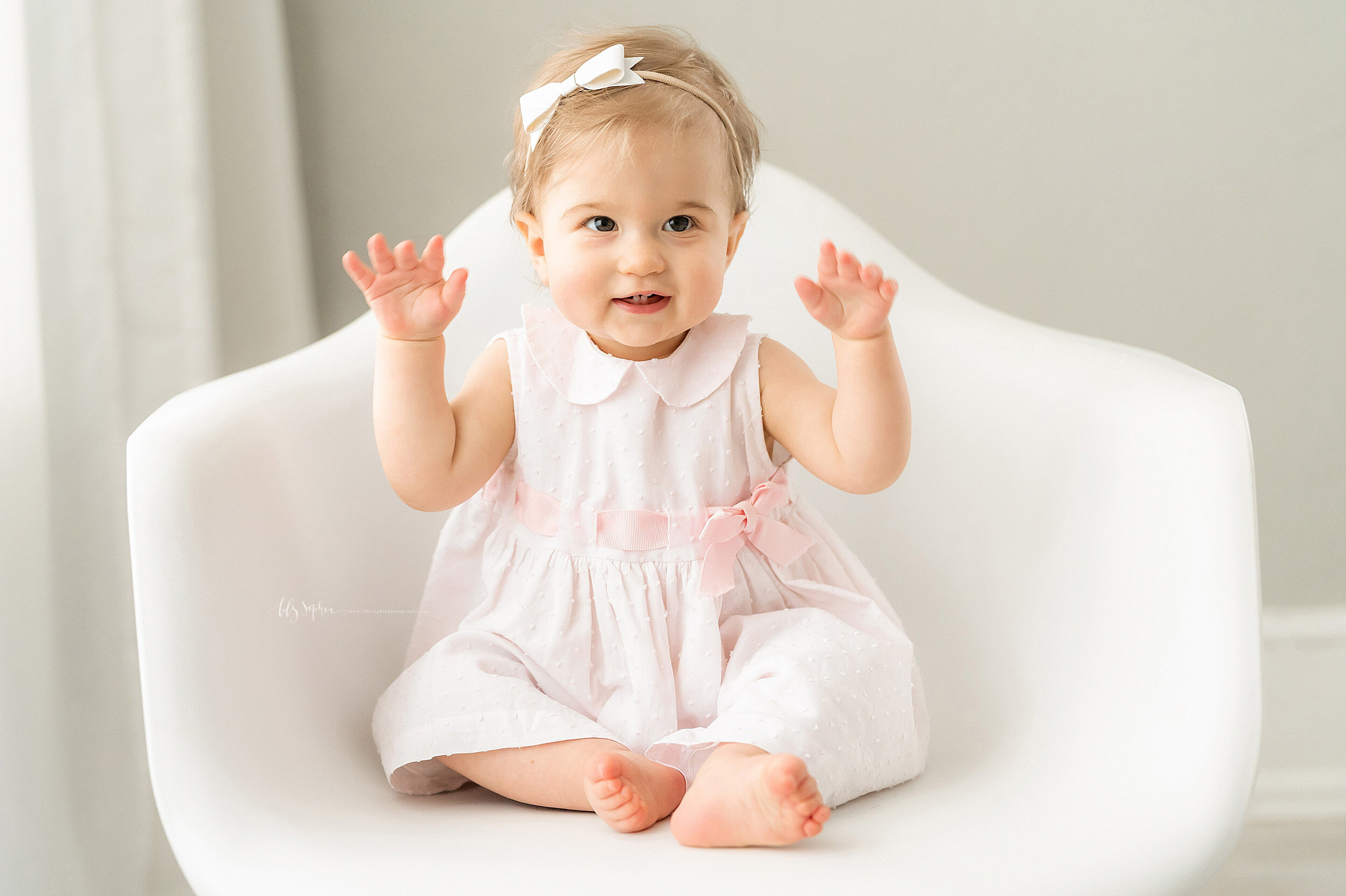  Milestone photo of a one year old girl as she sits in a white molded chair with her hands in the air showing how big she is taken in natural light in a Buckhead studio outside of Atlanta. 