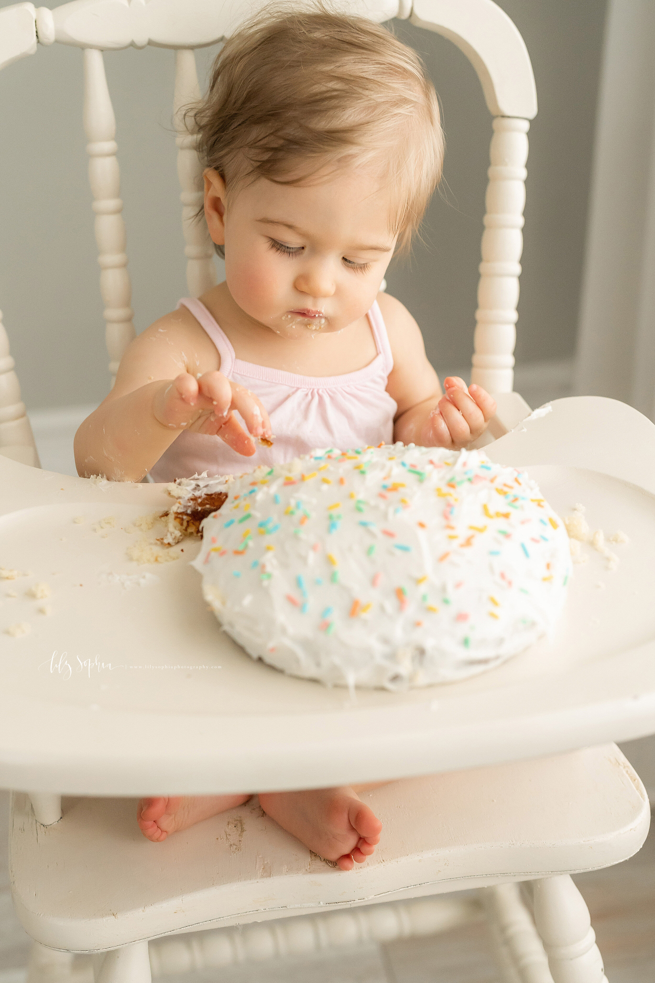  Photo of a one year old girl sitting in an antique high chair in natural light pensively deciding what part of her smash cake to taste taken in a studio in the Ansley area of Atlanta, Georgia. 