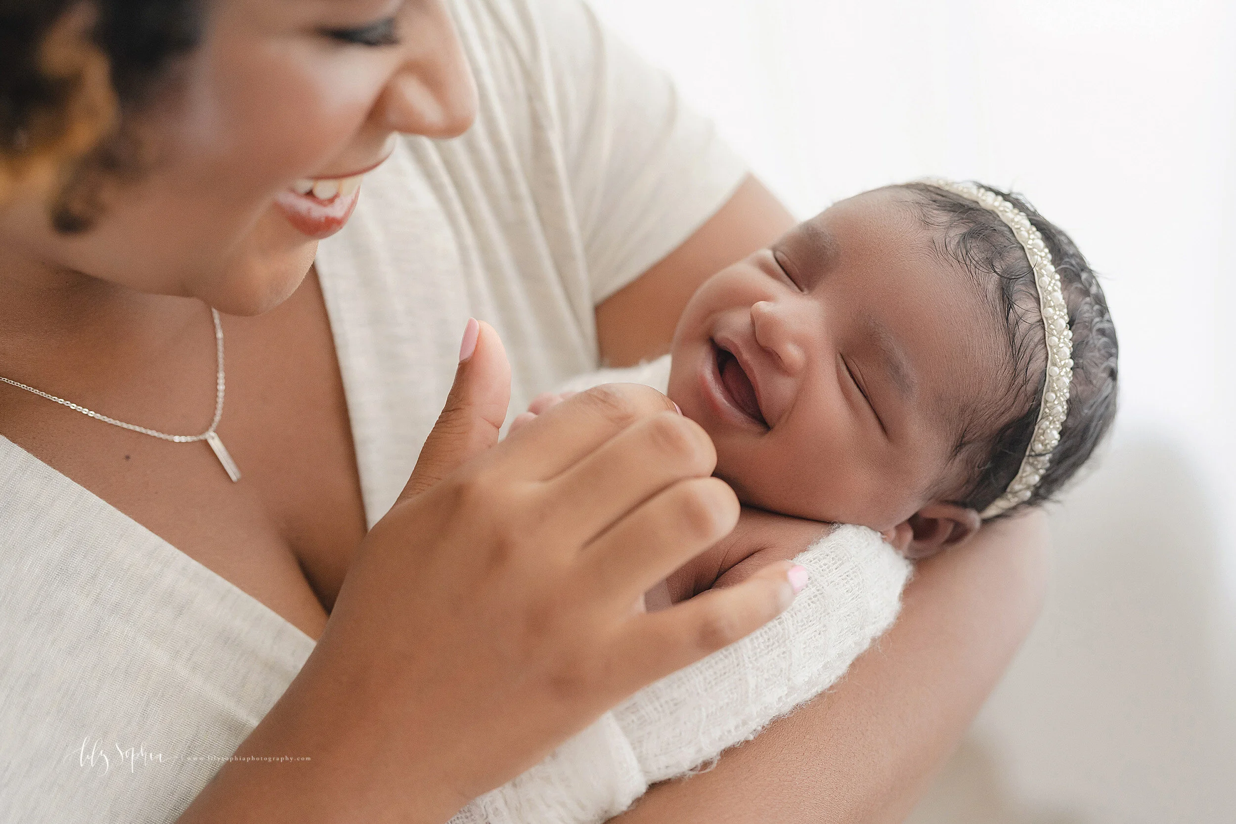 Newborn photo of an African-American mother holding her sleeping newborn daughter as she tickles her chin to get her to smile taken by Lily Sophia Photography in a natural light studio in the Poncey Highland area of Atlanta.