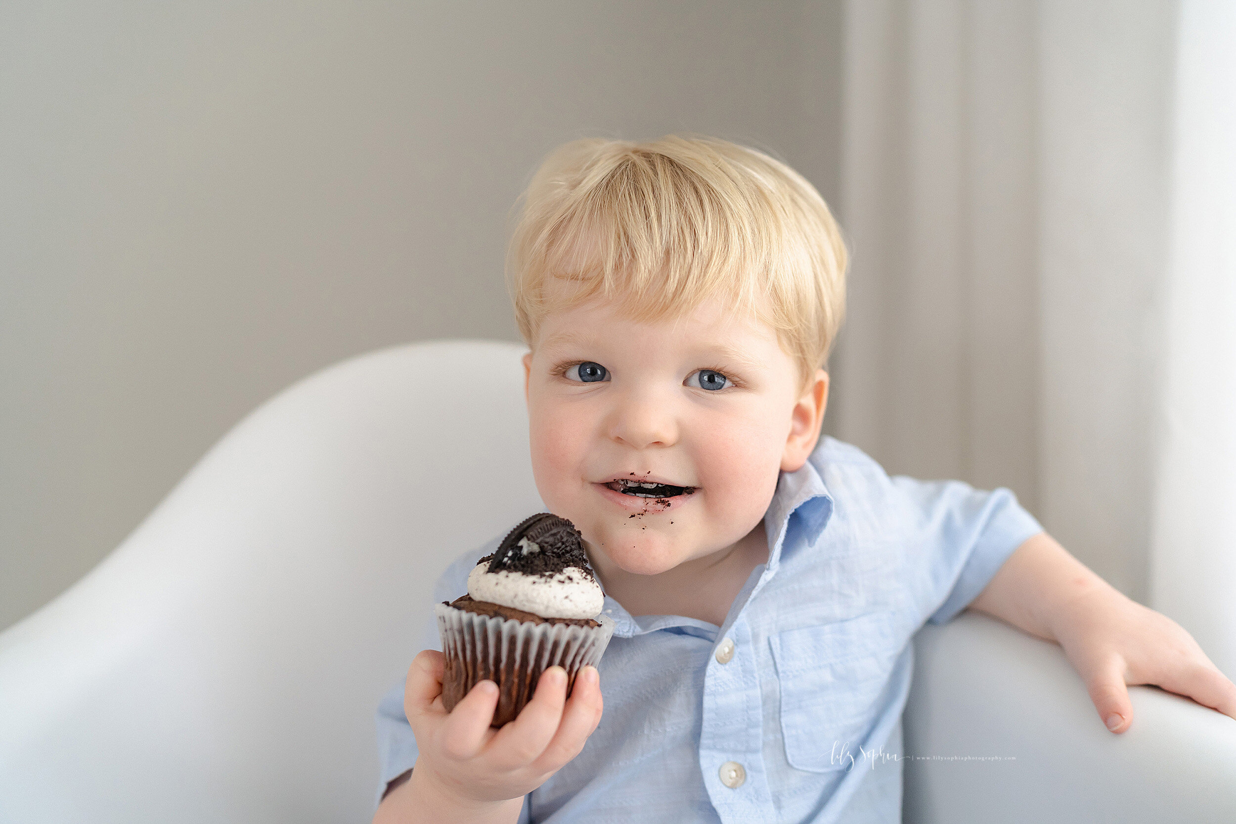  Photo of a blond haired, blue eyed two year old boy siting in a white molded chair holding a chocolate cupcake after taking a bite taken in a natural light studio near Buckhead in Atlanta, Georgia. 
