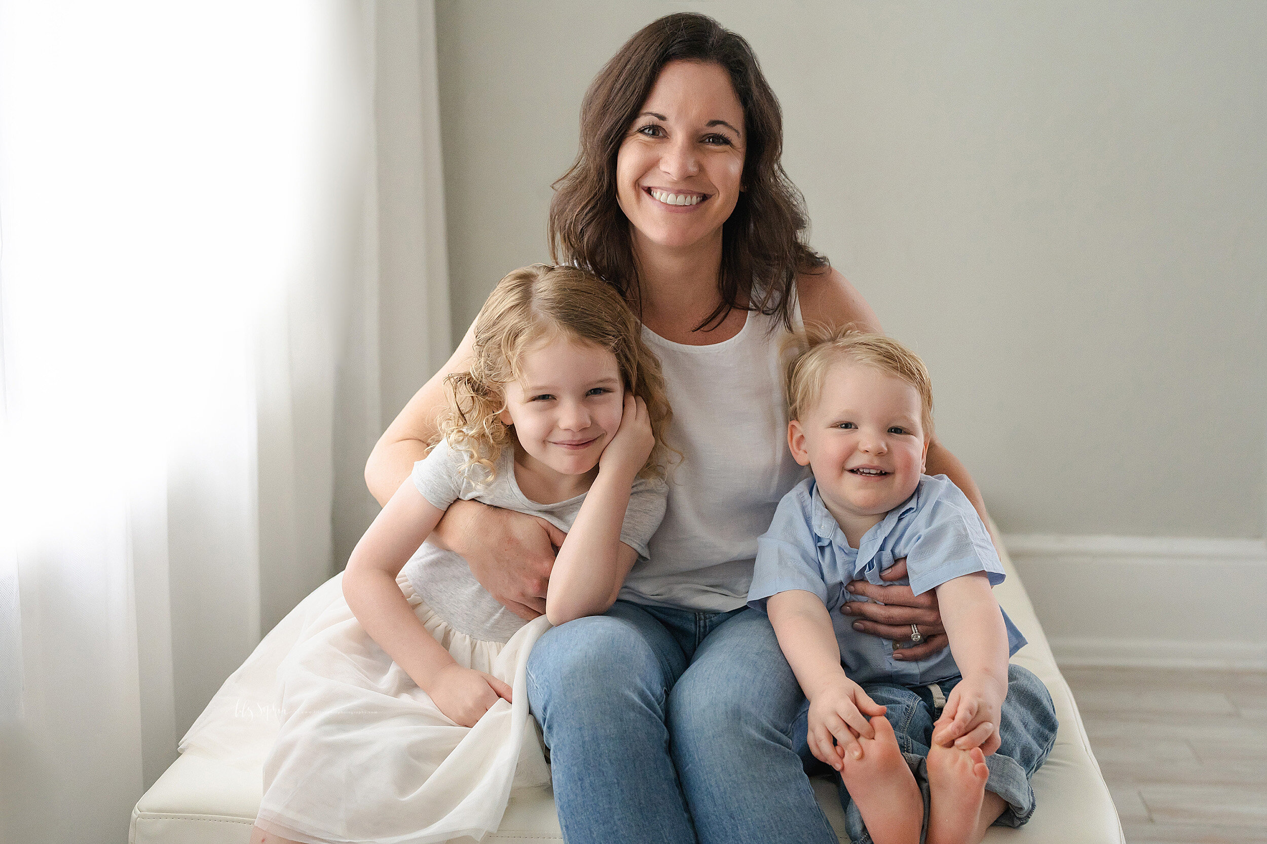  Family photo of a mom and her two children as they sit on a bench in front of window in natural light in an Atlanta studio near Virginia Highlands. 