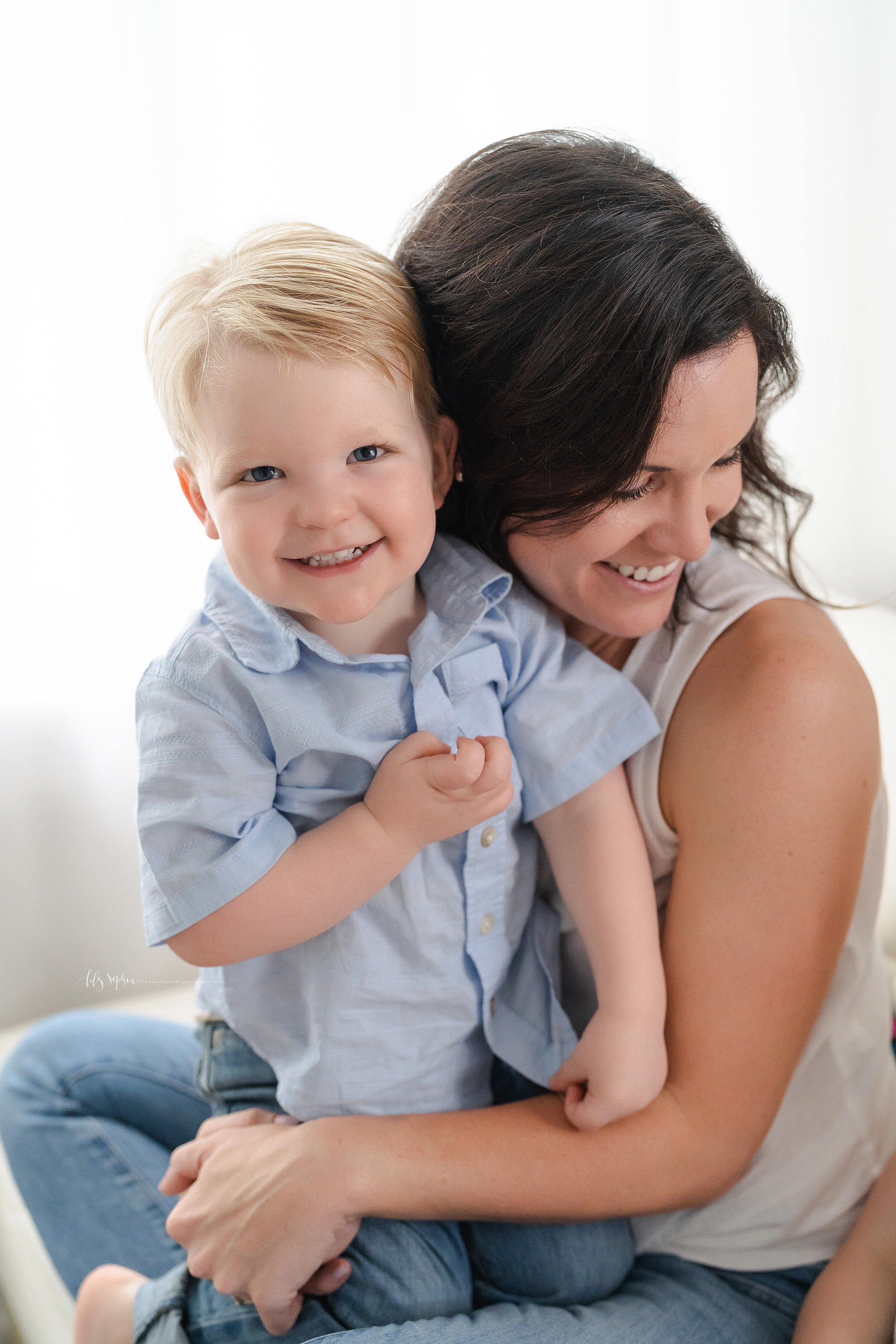  Family photo of a happy mom and her two year old son as she holds him on her lap and looks over her left shoulder while sitting in a chair in a studio in Poncey Highlands, Georgia in natural light. 
