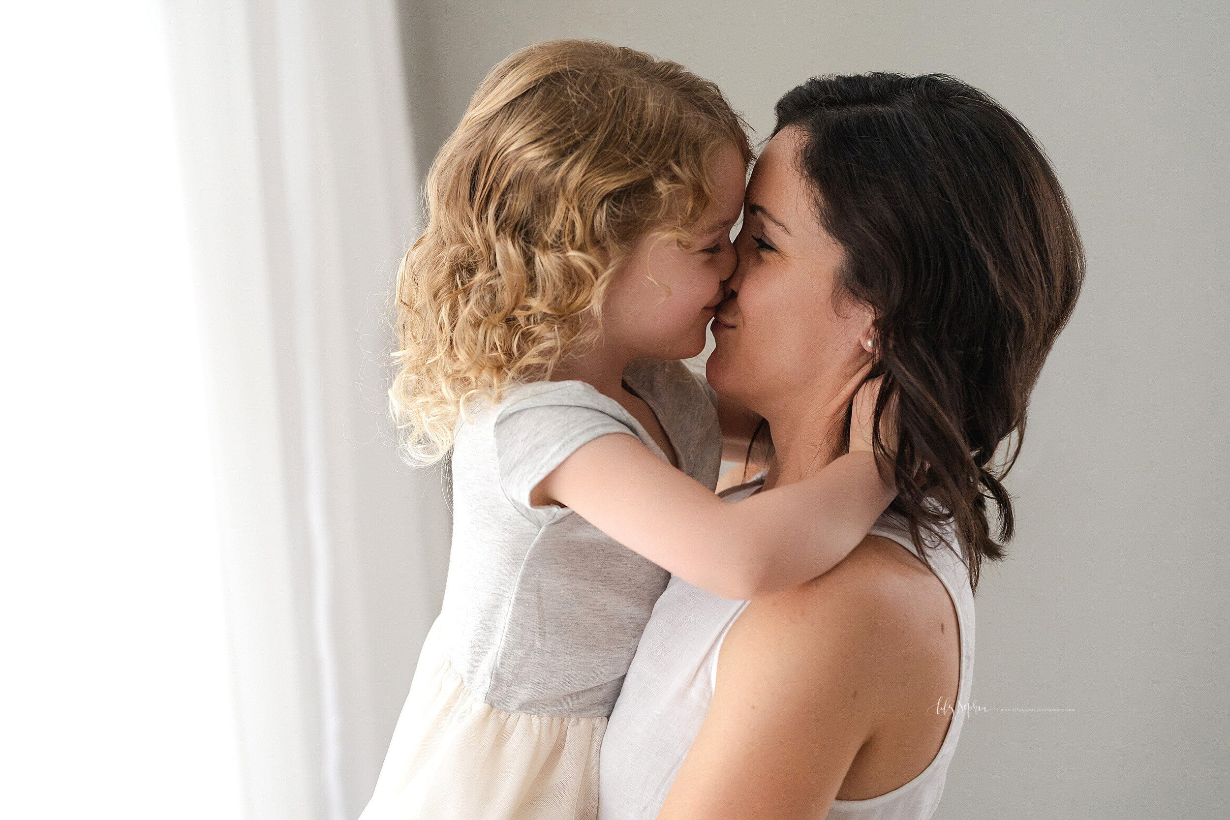  Photo of mom and her curly haired daughter as she stands in front of a window in natural light holding her daughter in her arms while her daughter wraps her hands around her mother’s neck and kisses her taken in a studio near Midtown in Atlanta, Geo
