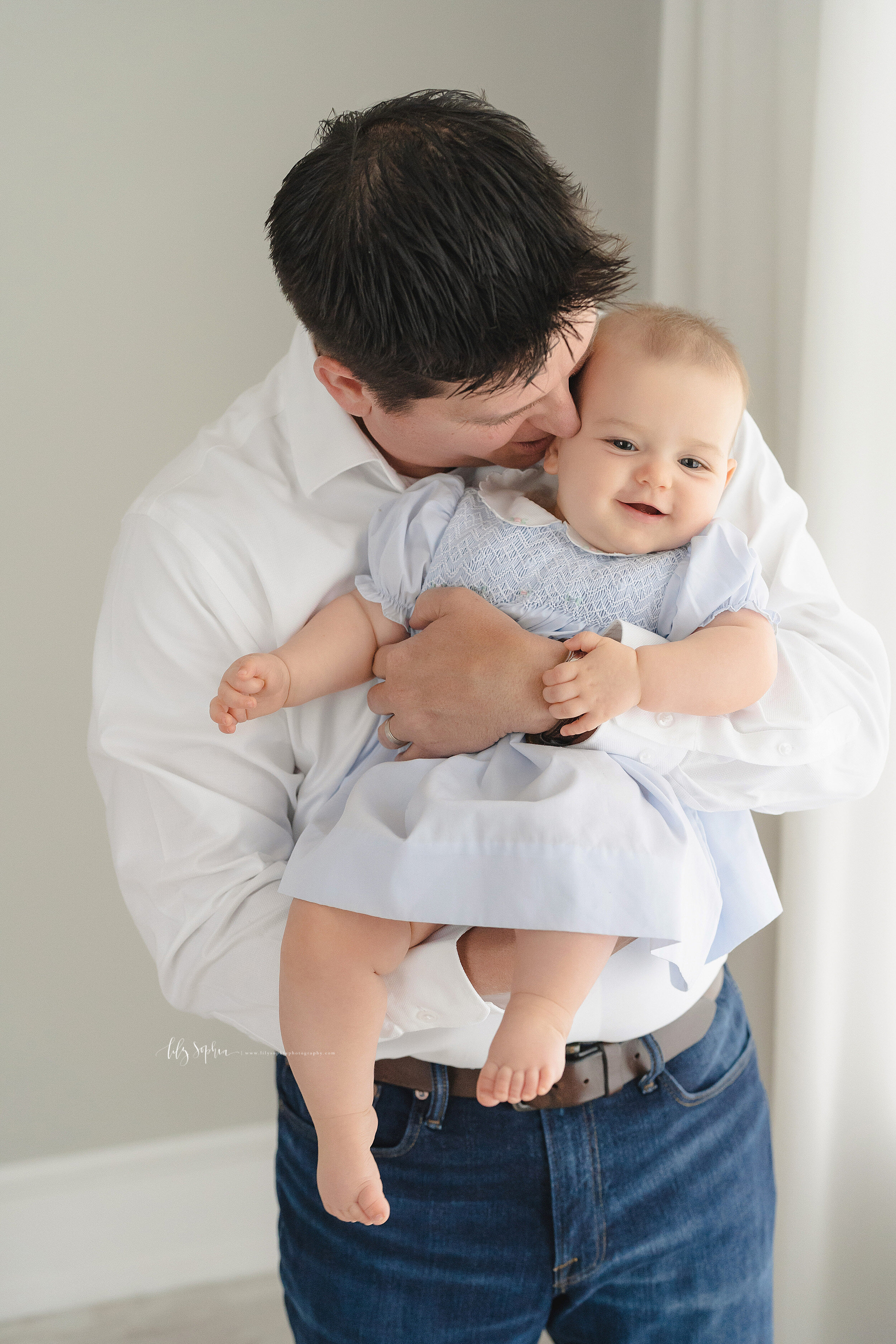  Family photo of a dad and his precious nine month old daughter as he whispers in her ear while holding her in front of his chest as he stands in natural light in an Atlanta studio. 