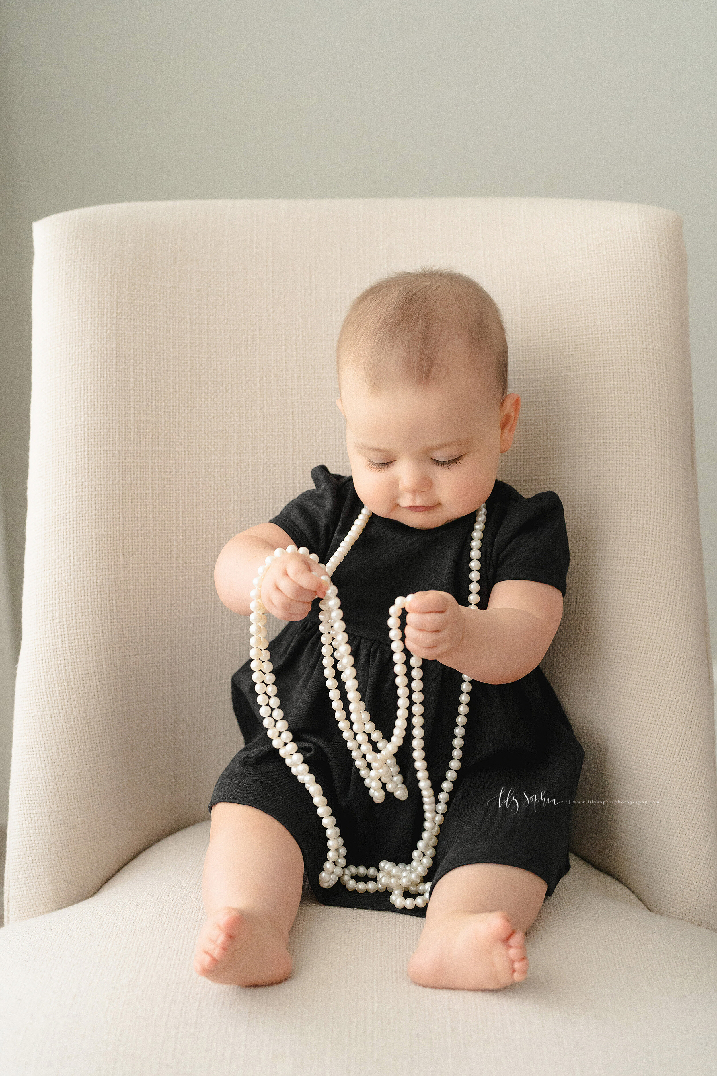  Precious photo of a nine month old girl sitting on a armless highback chair trying to figure out the long string of pearls around her neck taken in a studio near the Ansley Park area of Atlanta, Georgia in natural light. 