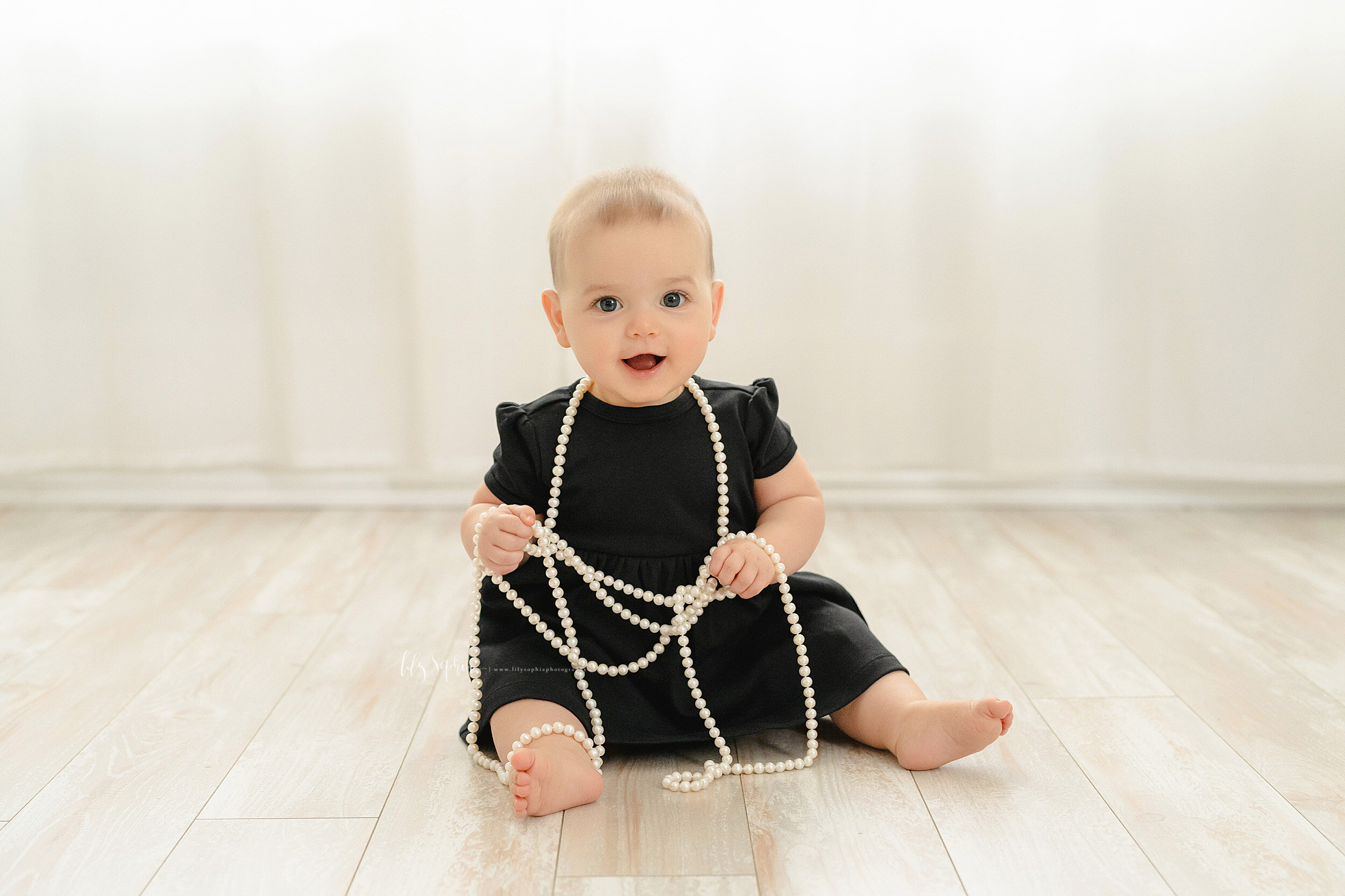  Photo of a smiling nine month old as she sits on the floor of a natural light studio and plays with her mother’s pearl necklace taken near the Old Fourth Ward area of Atlanta, Georgia. 
