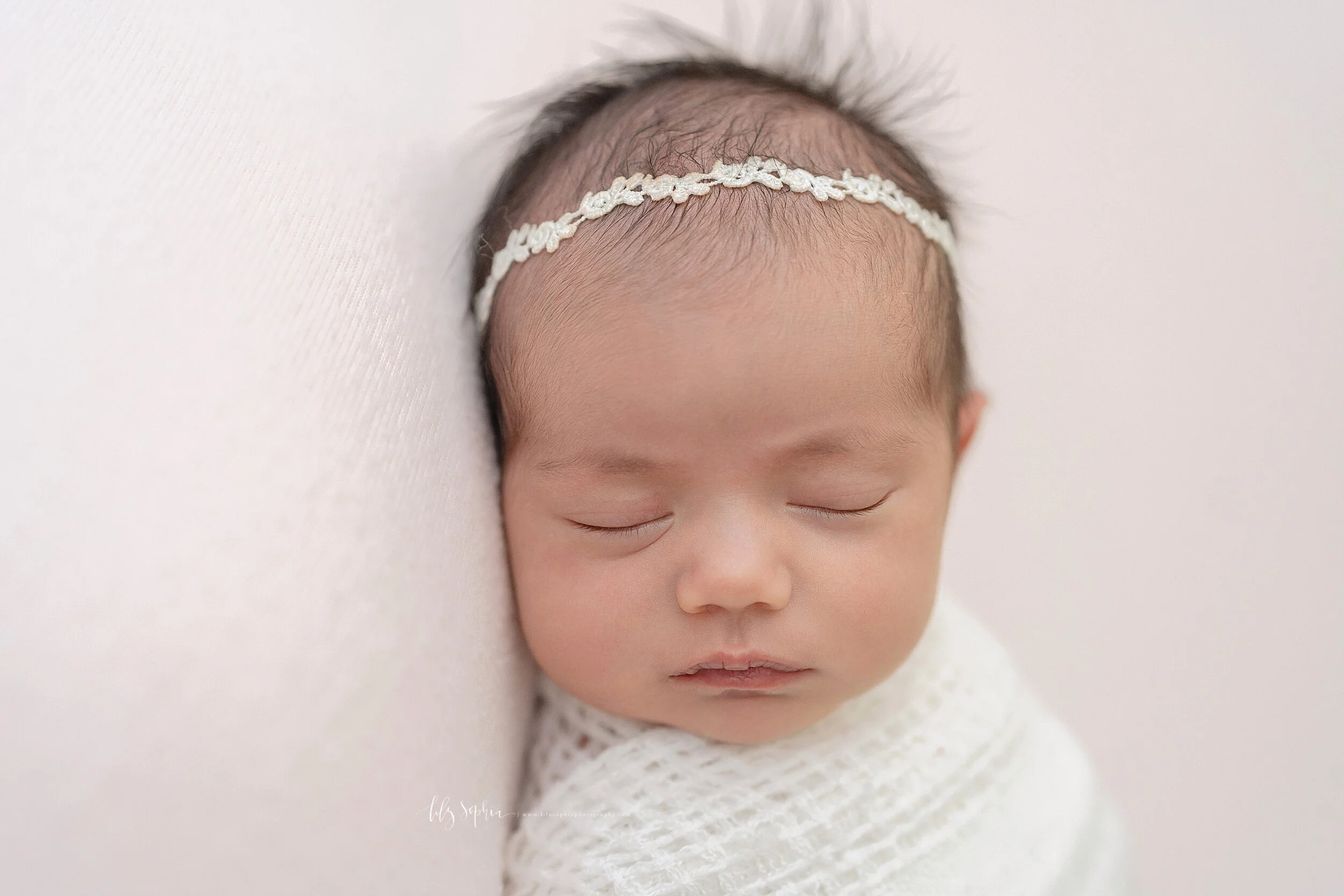  Photo of a newborn girl as she lies on her back with a soft white blanket swaddled to her chin and she wears a delicate woven headband in her hair as she contentedly sleeps in a studio in the Buckhead area of Atlanta in natural light. 