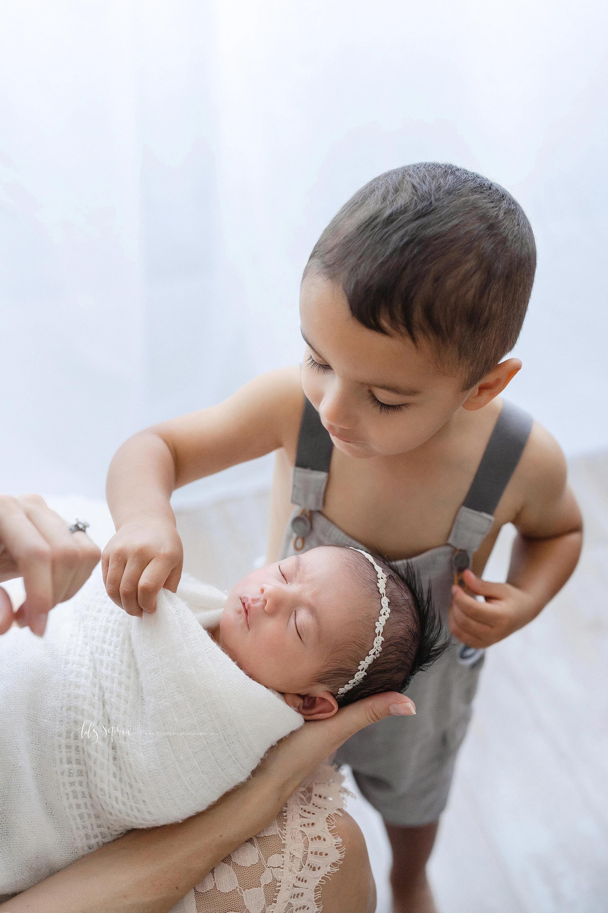  A curious big brother peeks inside the blanket swaddle of his newborn sister as she soundly sleeps on his mother’s lap in a studio near Poncey Highlands in Atlanta in natural light. 