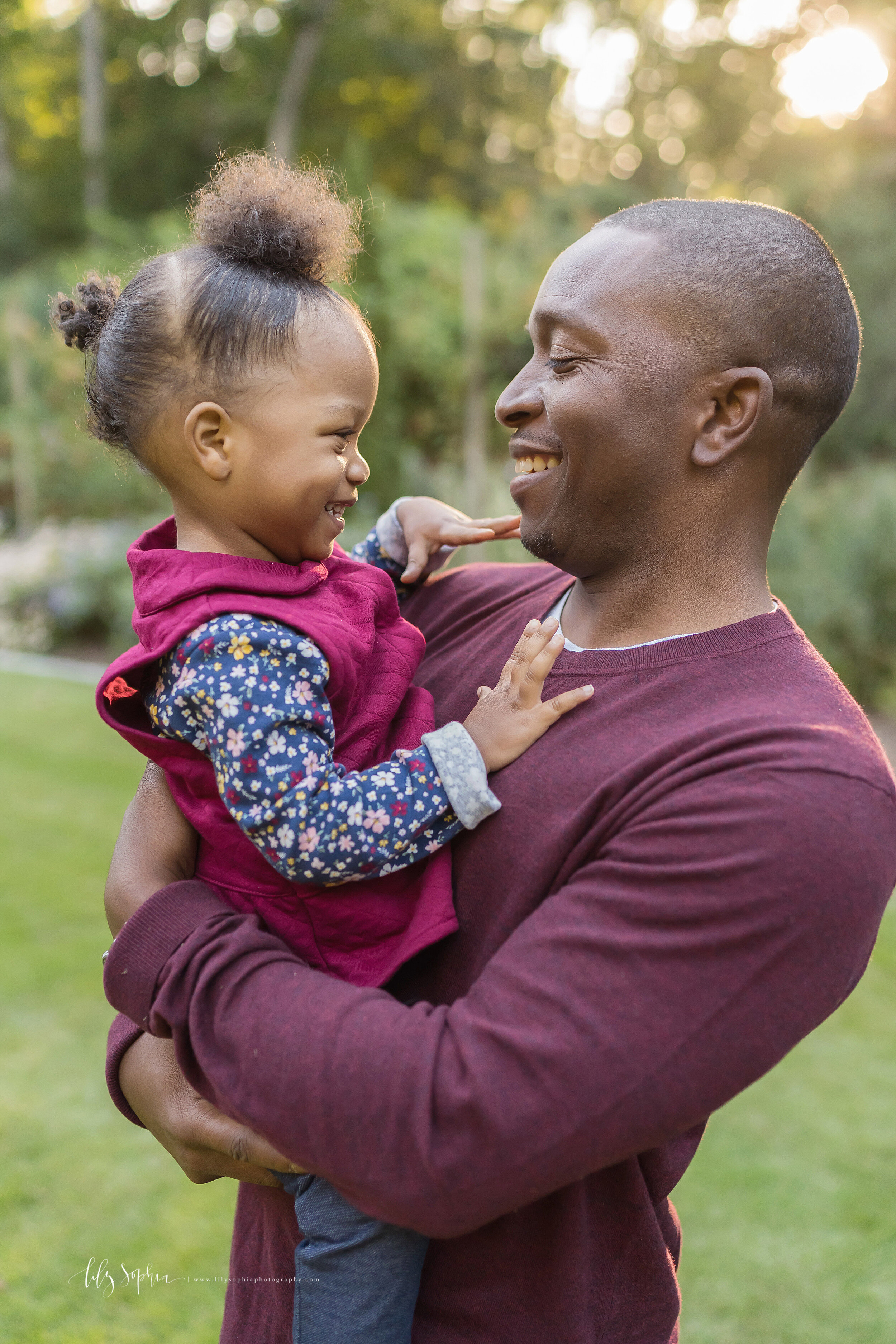  Family photo of a smiling African-American father as he holds his playful toddler daughter in his arms in a garden near Atlanta at sunset. 