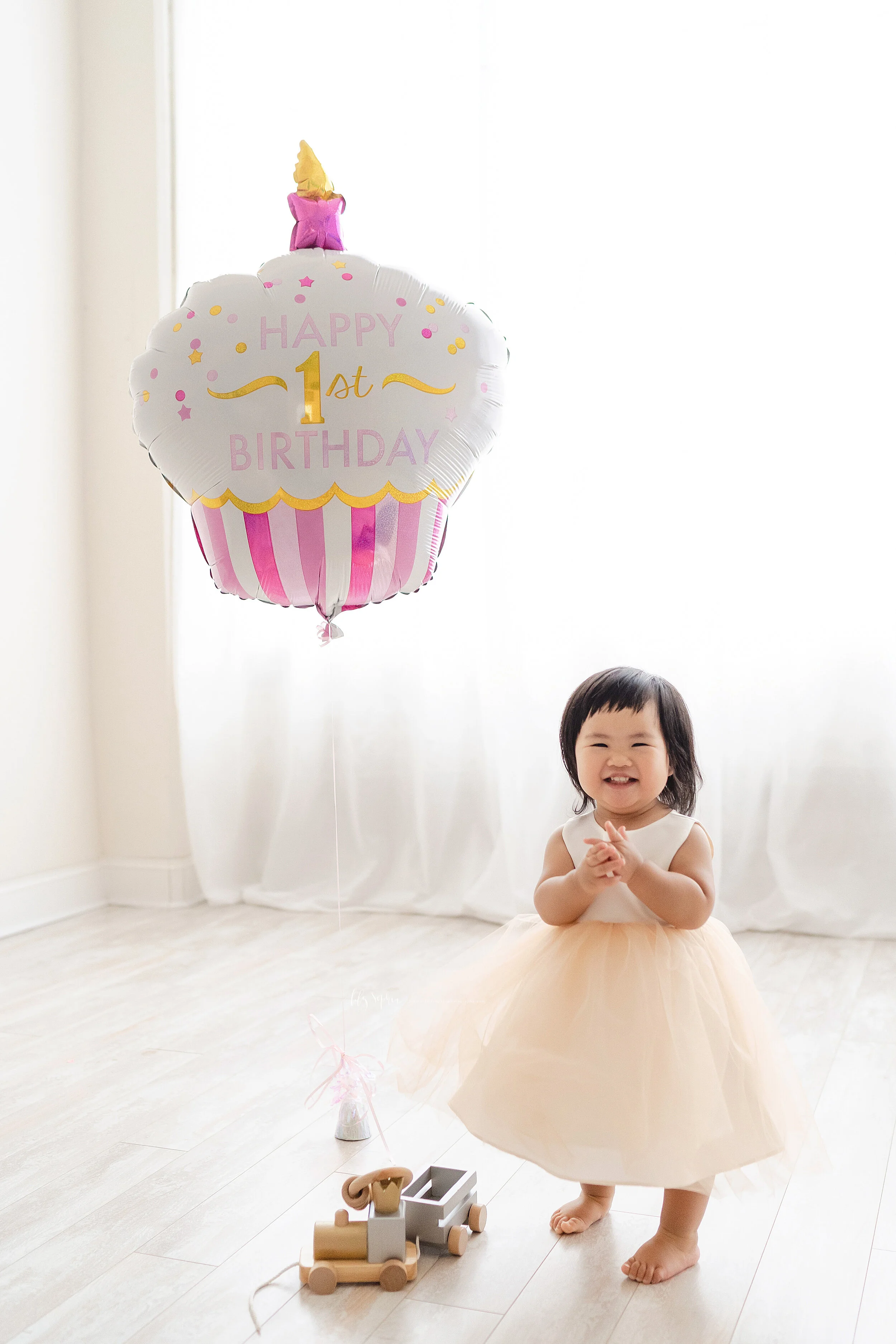 Milestone photo of an excited Asian one year old as she stands next to her birthday balloon and claps her hands in a Ansley Park studio in natural light.