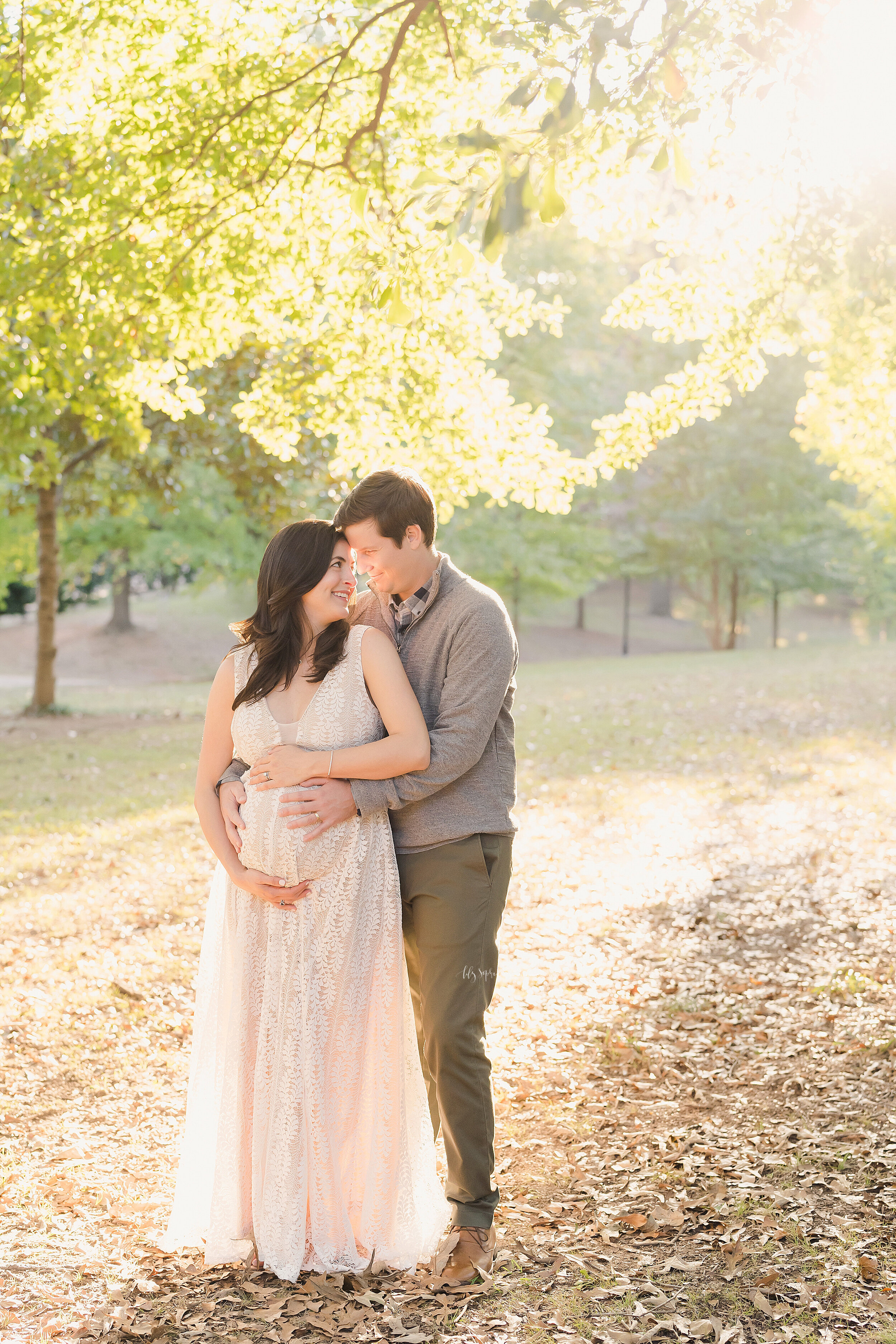  Maternity photo of a husband and wife as they stand and look lovingly at one another while they cherish their expectant child with the glow of the sunset surrounding them in a park in Atlanta. 