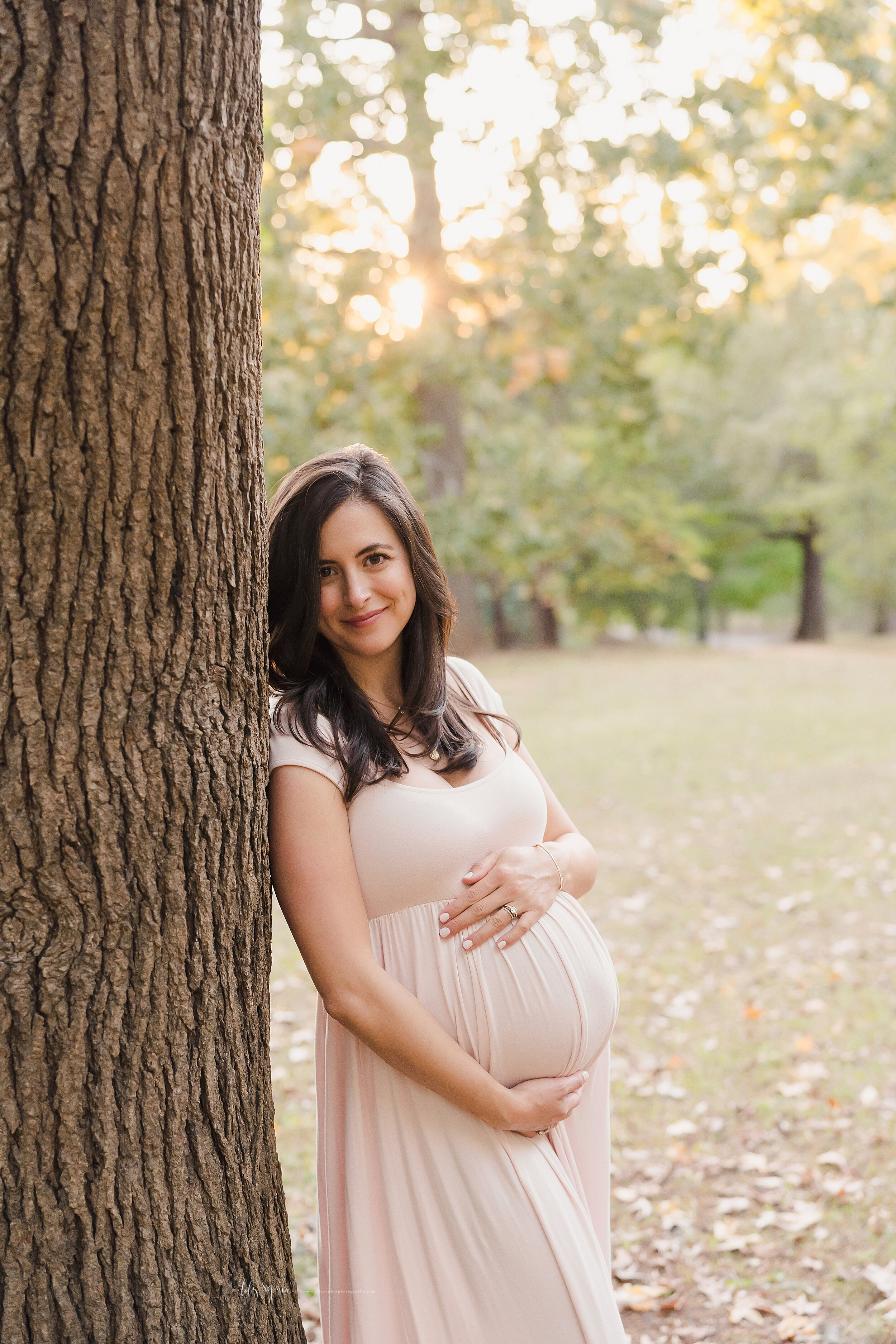  Maternity photo of a radiant pregnant woman as she stands against a tree and holds her left hand on top of her belly and her right hand below her belly in a park at sunset. 