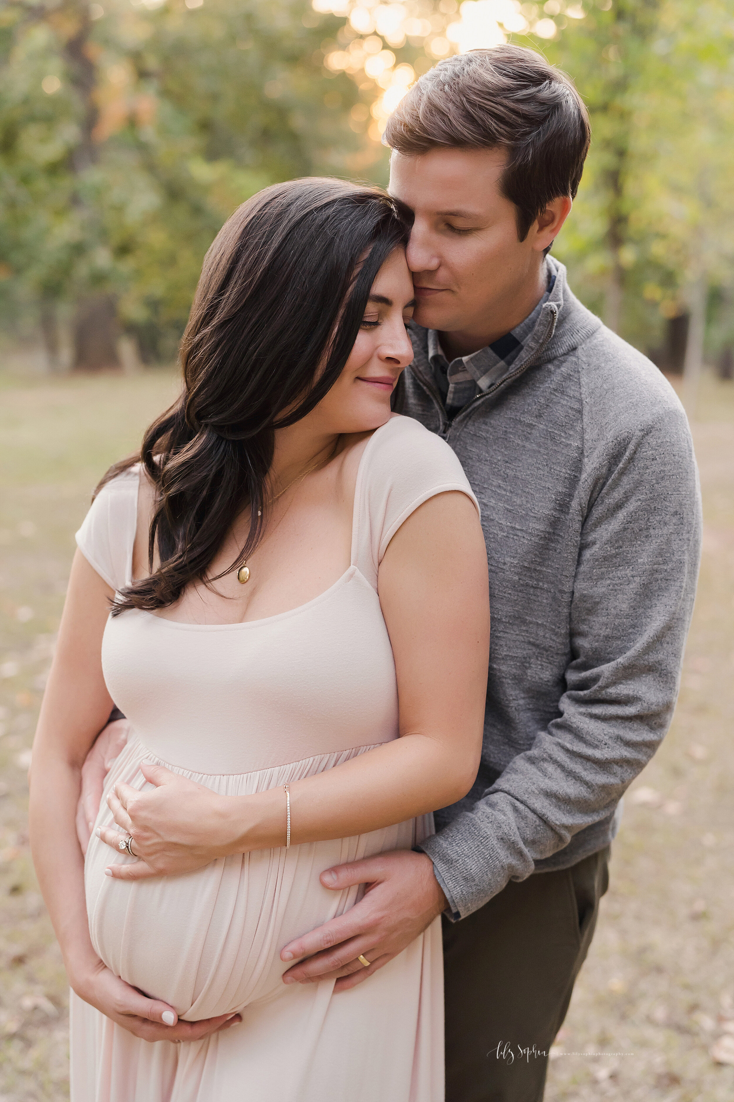  Maternity photo of a husband and wife as they stand in a park at sunset with the husband standing behind his wife and wrapping his hands around her to touch their child in utero and placing his head against his wife’s forehead as they treasure this 