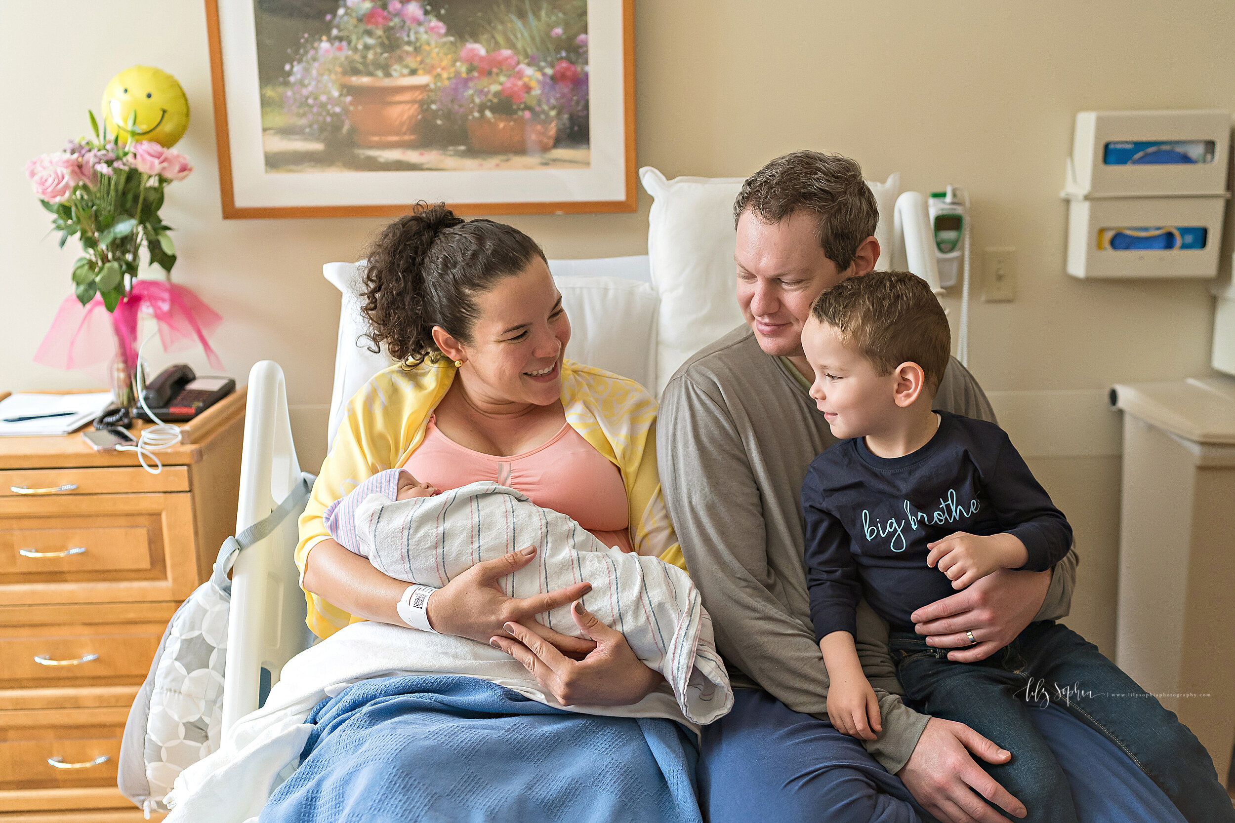  Family newborn photo of mom holding her daughter as she sits in her hospital bed next to her husband who is holding their toddler son on his lap in his big brother t-shirt as he gets introduced to his baby sister in an Atlanta hospital taken with na