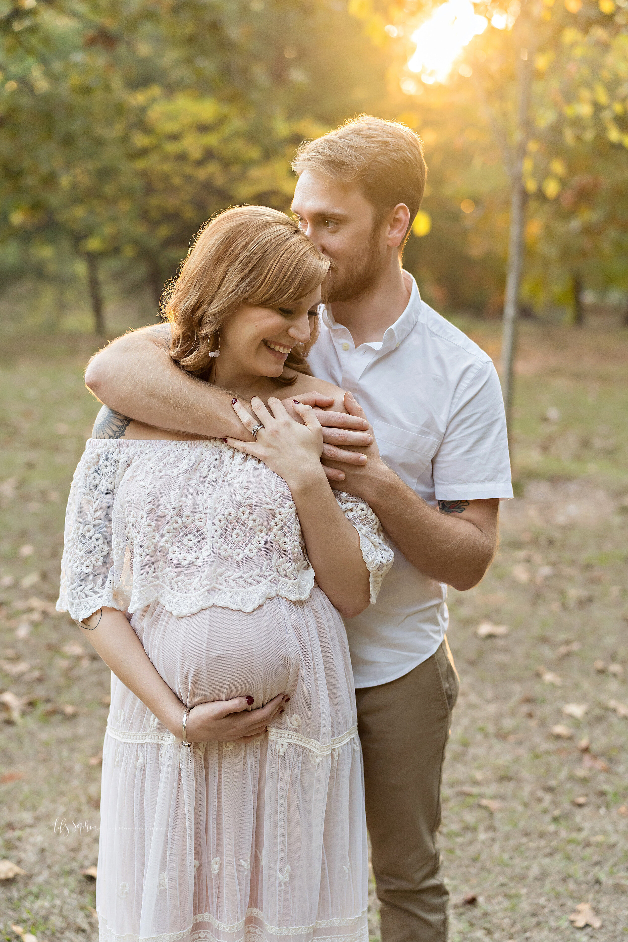  Maternity photo shoot of a husband and wife as the husband stands behind his wife with his arms around her shoulders and he whispers in her ear to make her laugh at sunset during autumn in an Atlanta park. 