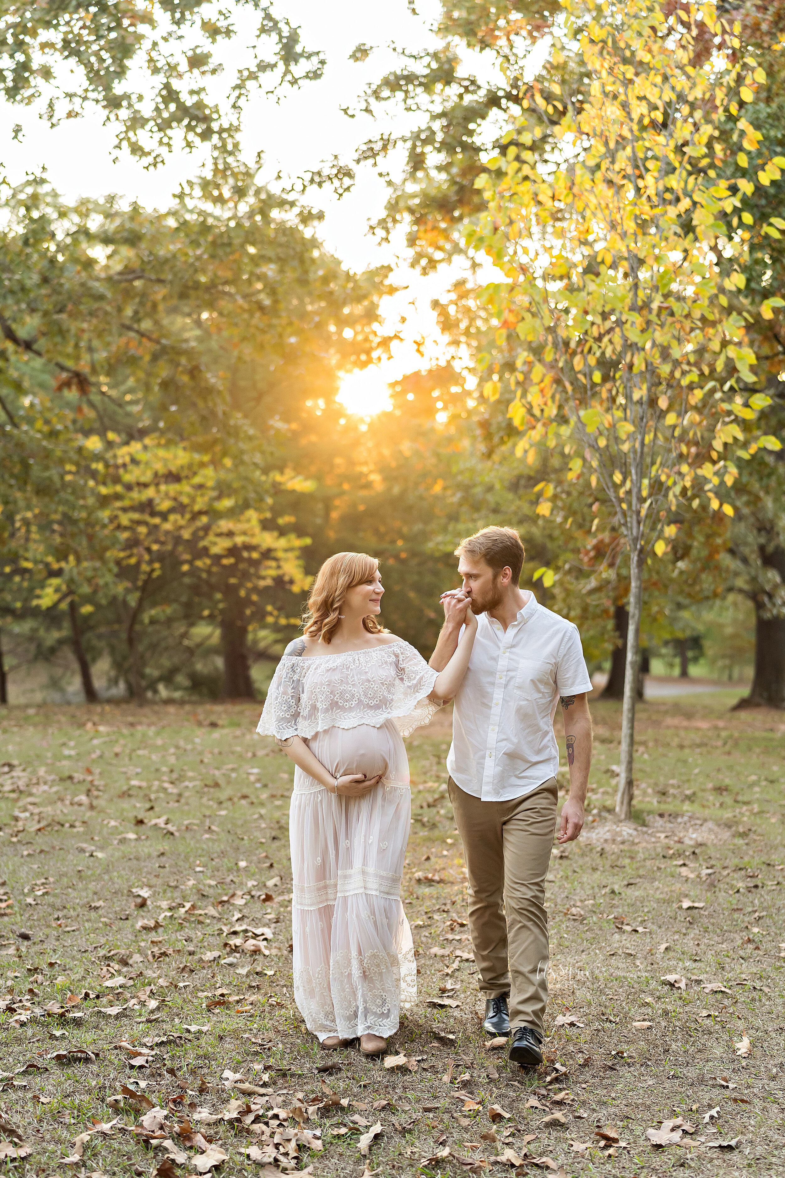  Maternity photograph of a husband and wife as they walk through an Atlanta park at sunset during autumn and the husband kisses his wife’s hand. 