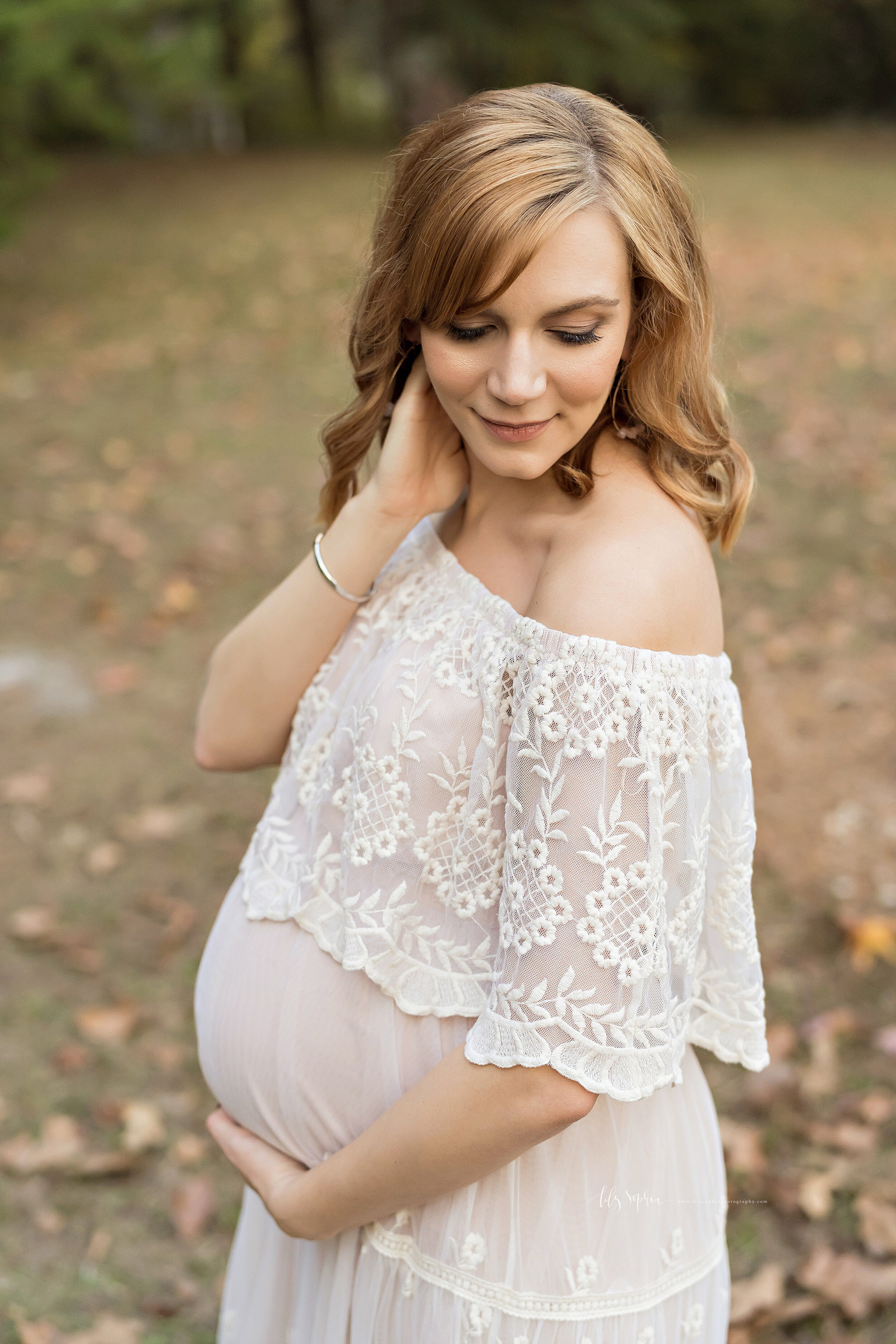  Maternity photo of a red-haired woman as she stands in a white off -the-shoulder dress in an Atlanta park  during autumn with her right hand on her neck and her left hand holding her child in utero as she contemplates the birth of her child. 