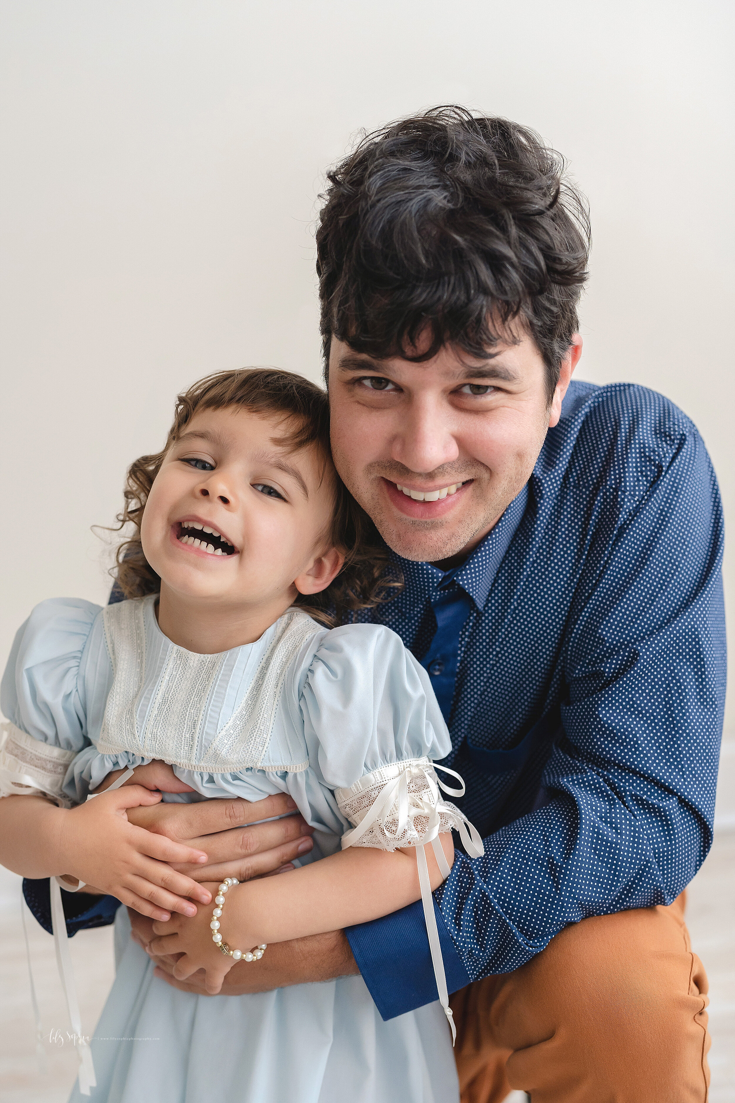  Family photo of a happy two year old with her dad as he kneels down and squeezes his daughter close to him while holding her around her waist in a studio Poncey-Highlands in natural light. 