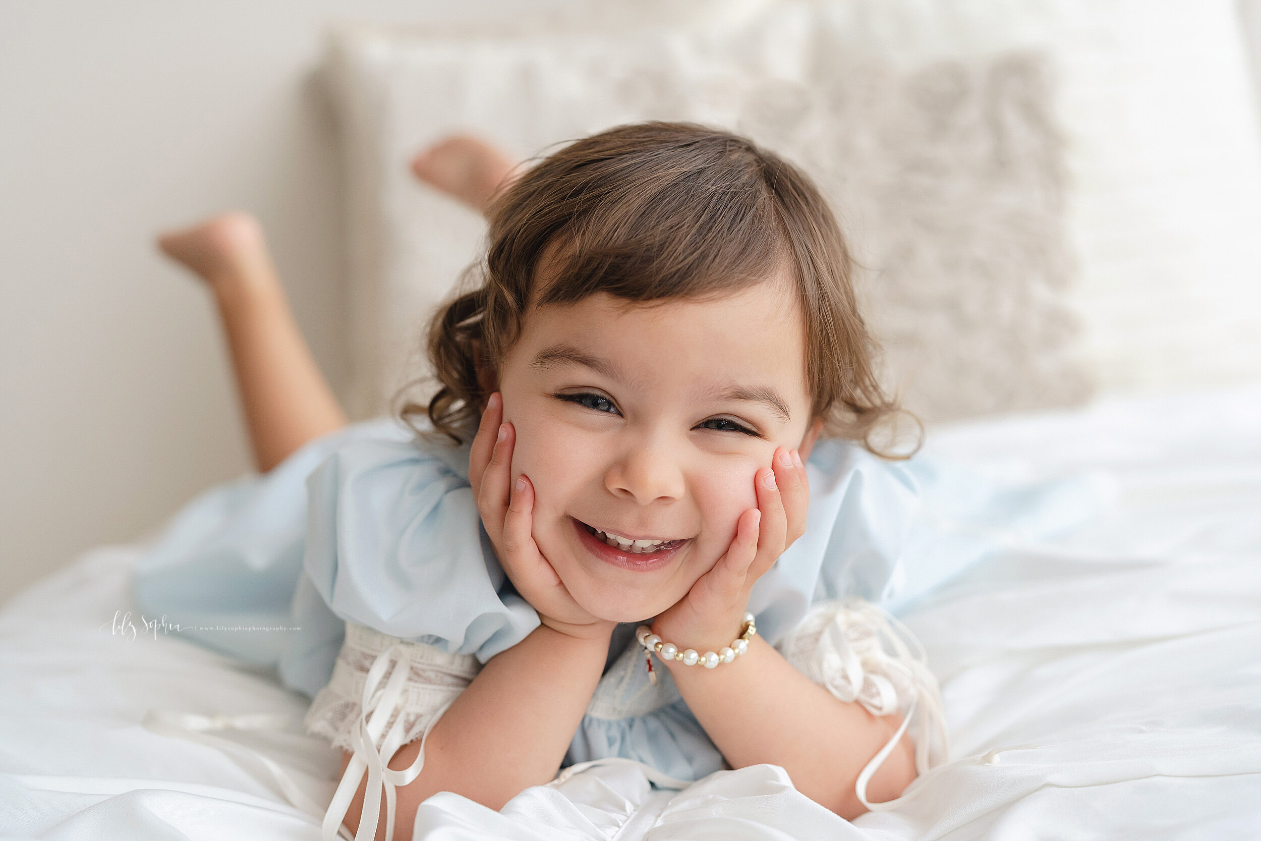  Milestone photo of a smiling two year old as she lies on her stomach on a bed in natural light with her hands on her cheeks in a studio near Inman Park in Atlanta. 