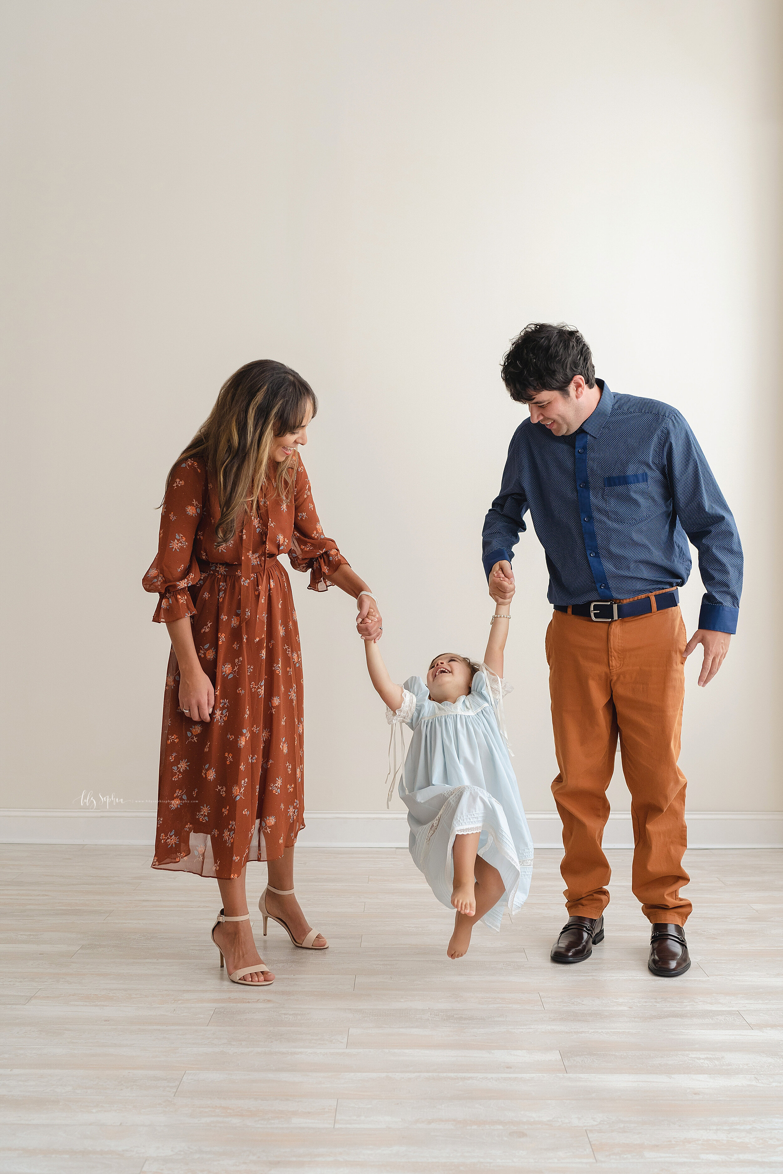  Family photo with a two-year old girl as she holds hands with her mom and dad in a natural light studio in the Buckhead area of Atlanta and her parents swing her between them. 