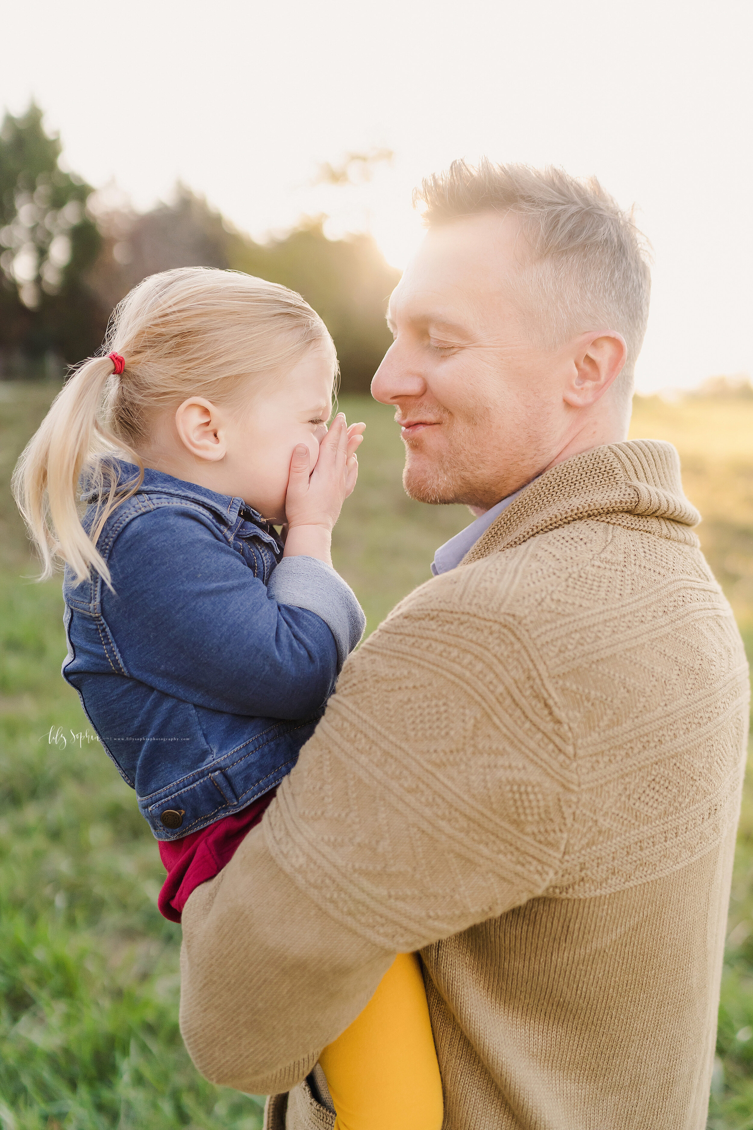  Family photo of a toddler daughter as she covers her face with her hands and giggles as her dad who is holding her, tries to kiss her with the sun setting in the background in a field near Atlanta, Georgia. 
