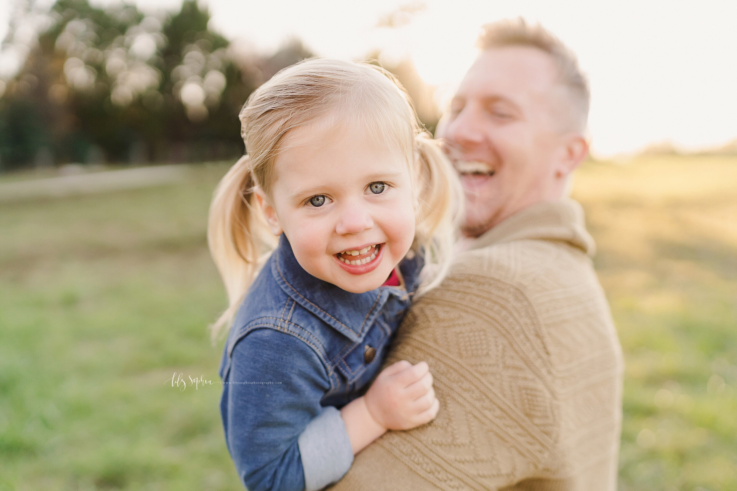  Family photo of a smiling blond haired, blue eyed little girl with pig tails in her hair as she is held in her dad’s arms in a field at sunset in Atlanta, Georgia. 