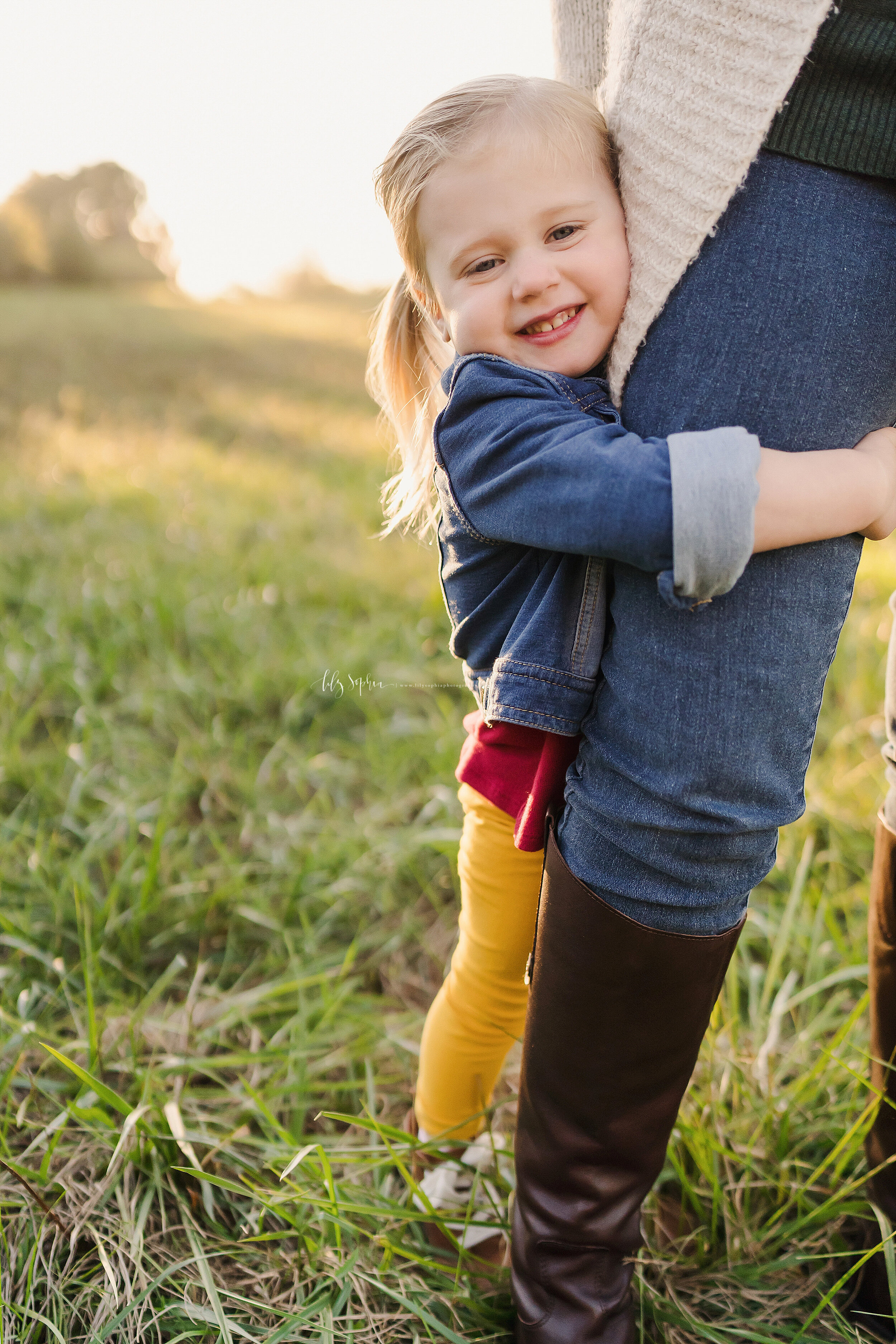  Family photo of a smiling toddler girl hugging her mother’s leg as they stand in a field at sunset near Atlanta, Georgia. 