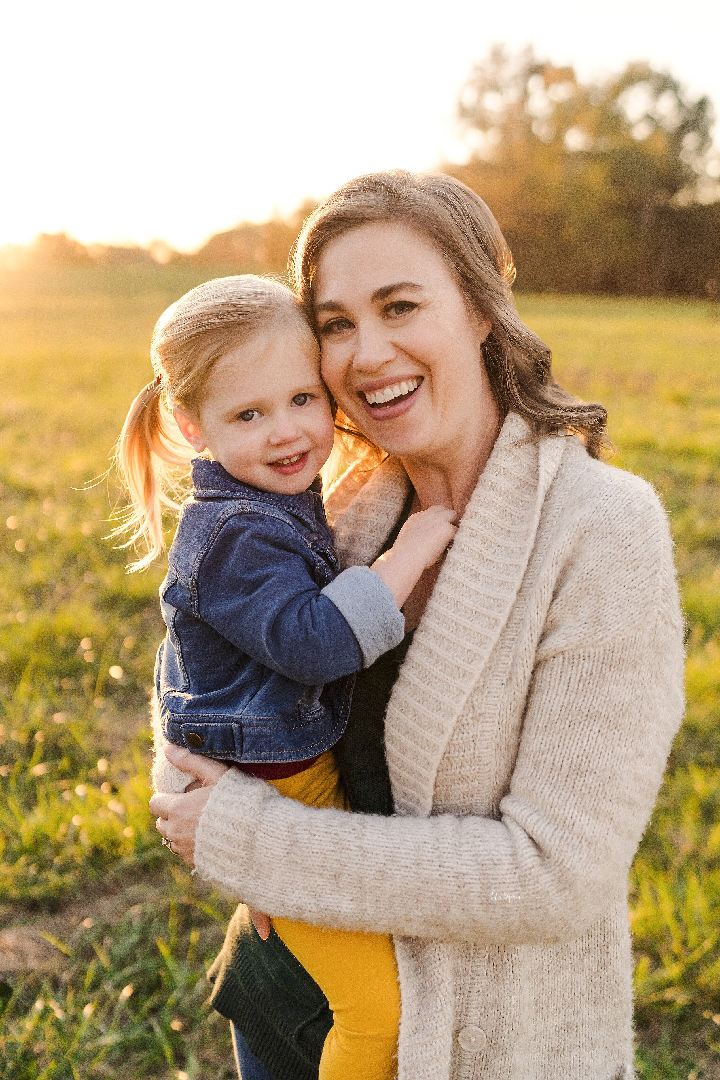  Family photography in an Atlanta field at sunset of a happy mother holding her toddler daughter as mom places her cheek against her daughter’s forehead. 