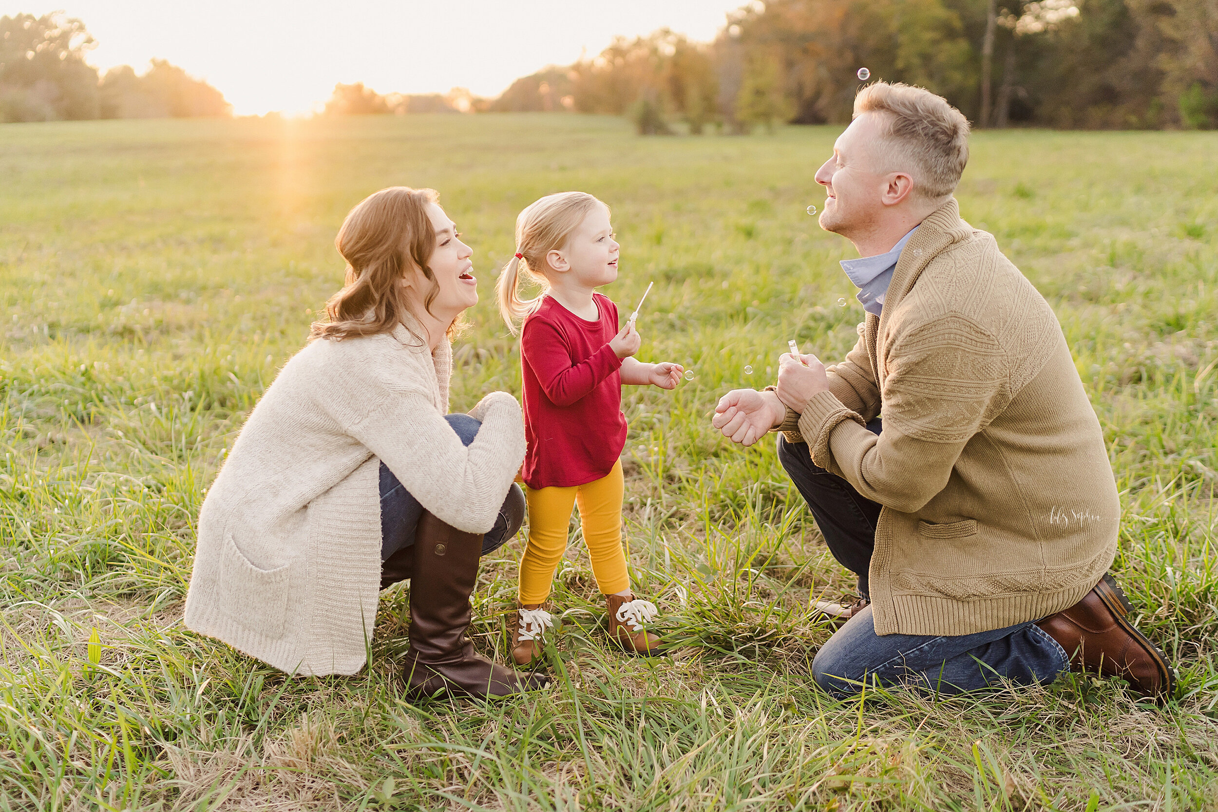  Family photo of three as mom crouches in a field near Atlanta with her daughter in front of her blowing bubbles at her husband who is kneeling and facing the toddler daughter and her mom at sunset. 