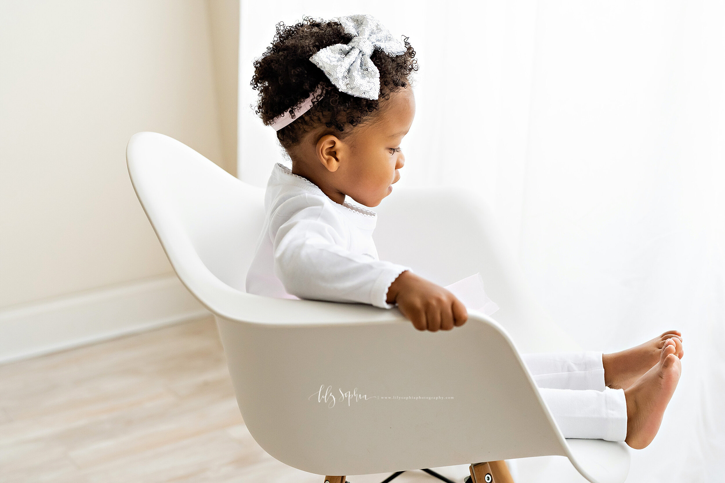  Milestone photo of a serious African-American two year old as she sits in a white molded chair and looks at her bare feet in front of a large natural light window in a studio in the Midtown area of Atlanta, Georgia. 