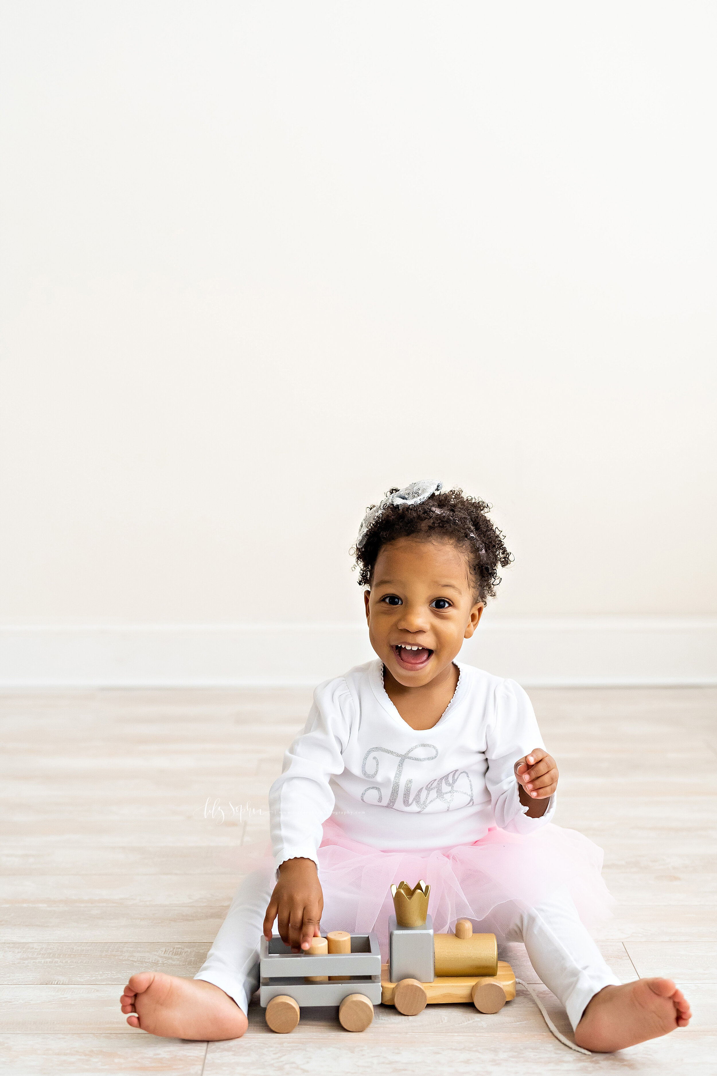  Milestone photo of an excited two year old girl as she sits on the floor of a natural light studio in the Virginia Highlands area of Atlanta sporting a white, long sleeved T-Shirt with a silver cursive Two on the front, white leggings and a pink tut