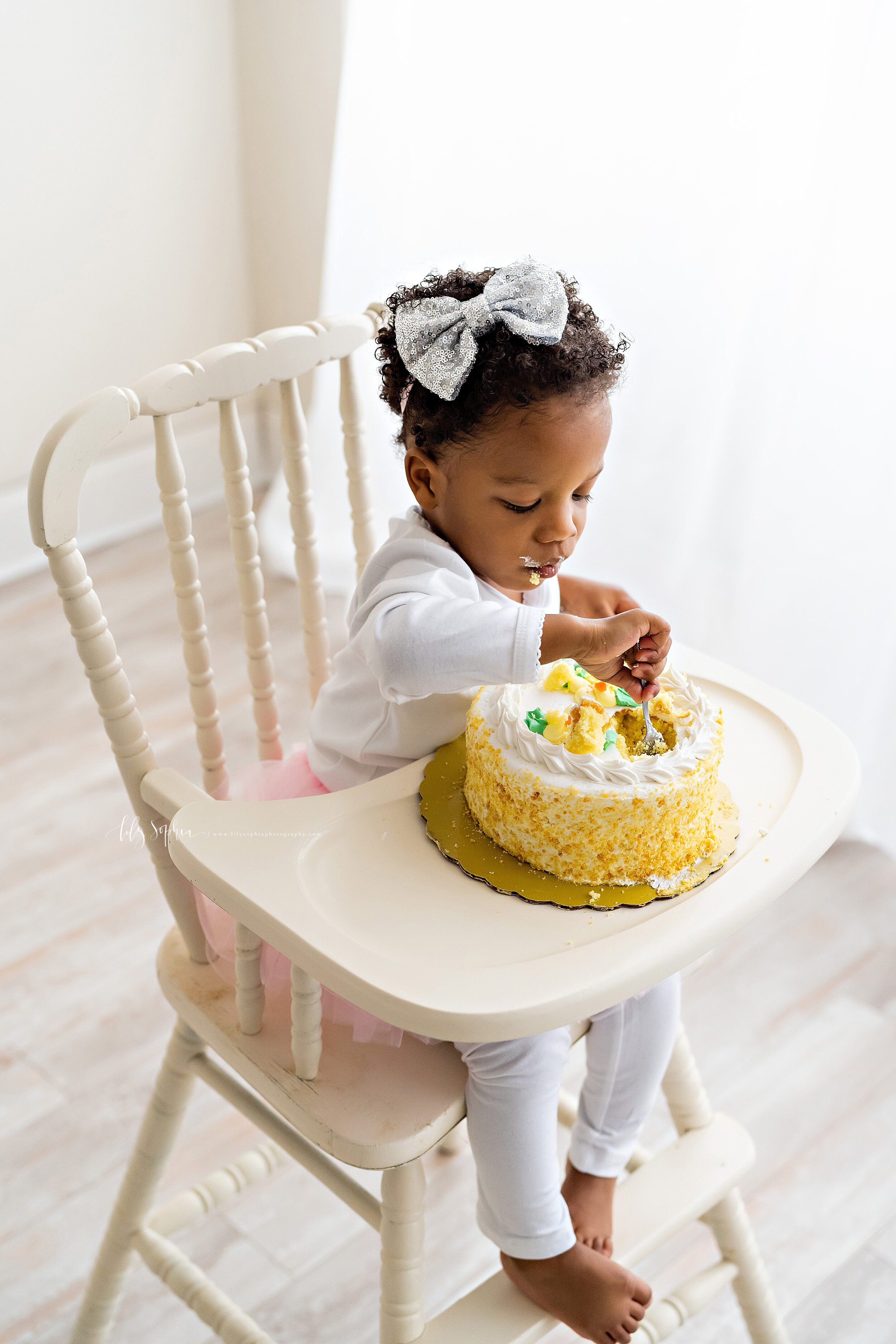  Milestone photograph of an African-American girl as she digs into her birthday cake with a spoon while sitting in a wooden antique high chair in front of a natural light window in a studio in the Peachtree Hills area of Atlanta, Georgia to celebrate