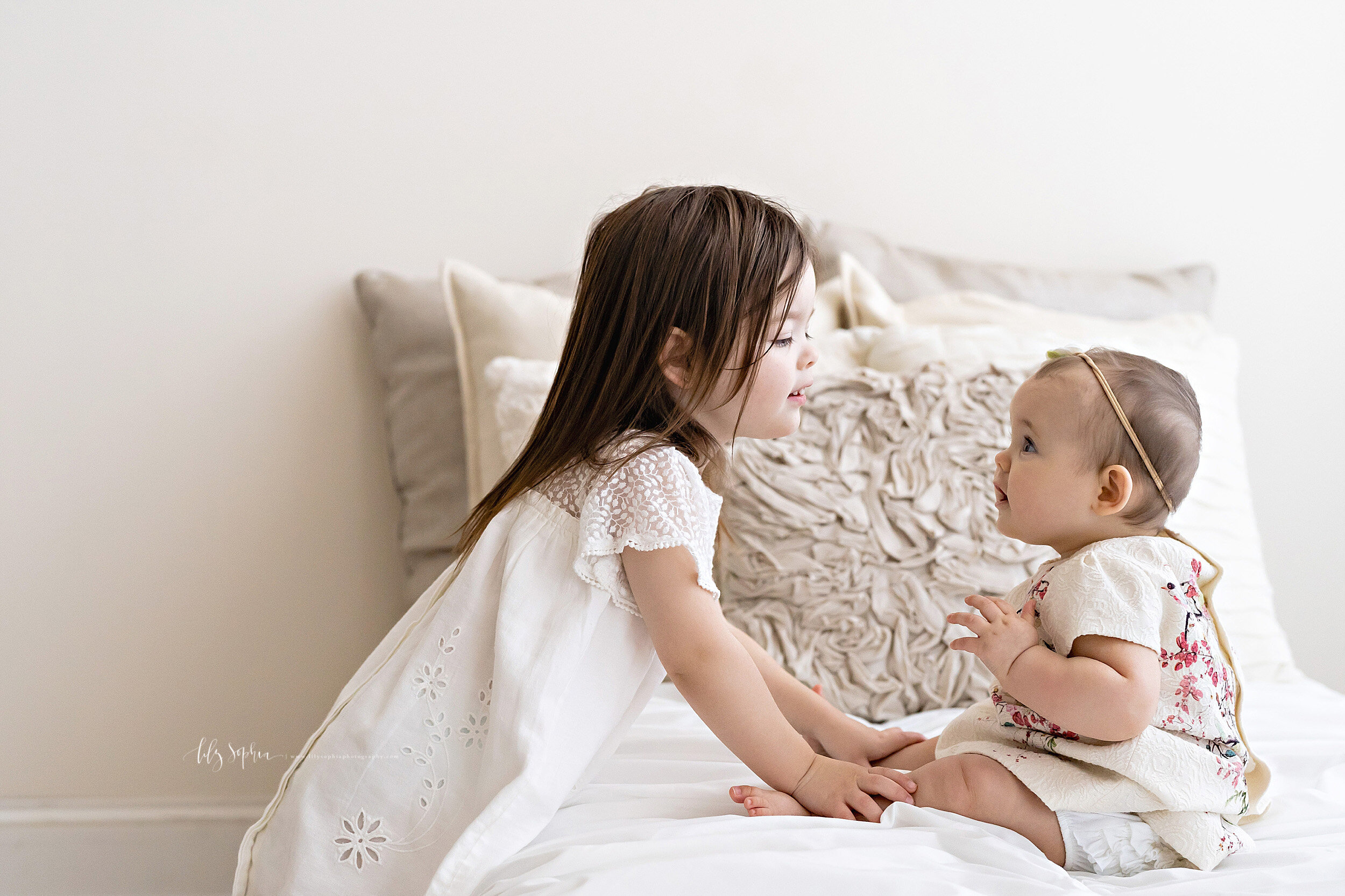  Family photo of a toddler girl and her baby sister as the baby sits on the bed and the toddler holds her sisters legs while they look at one another taken in natural light in a Virginia Highlands studio in Atlanta. 