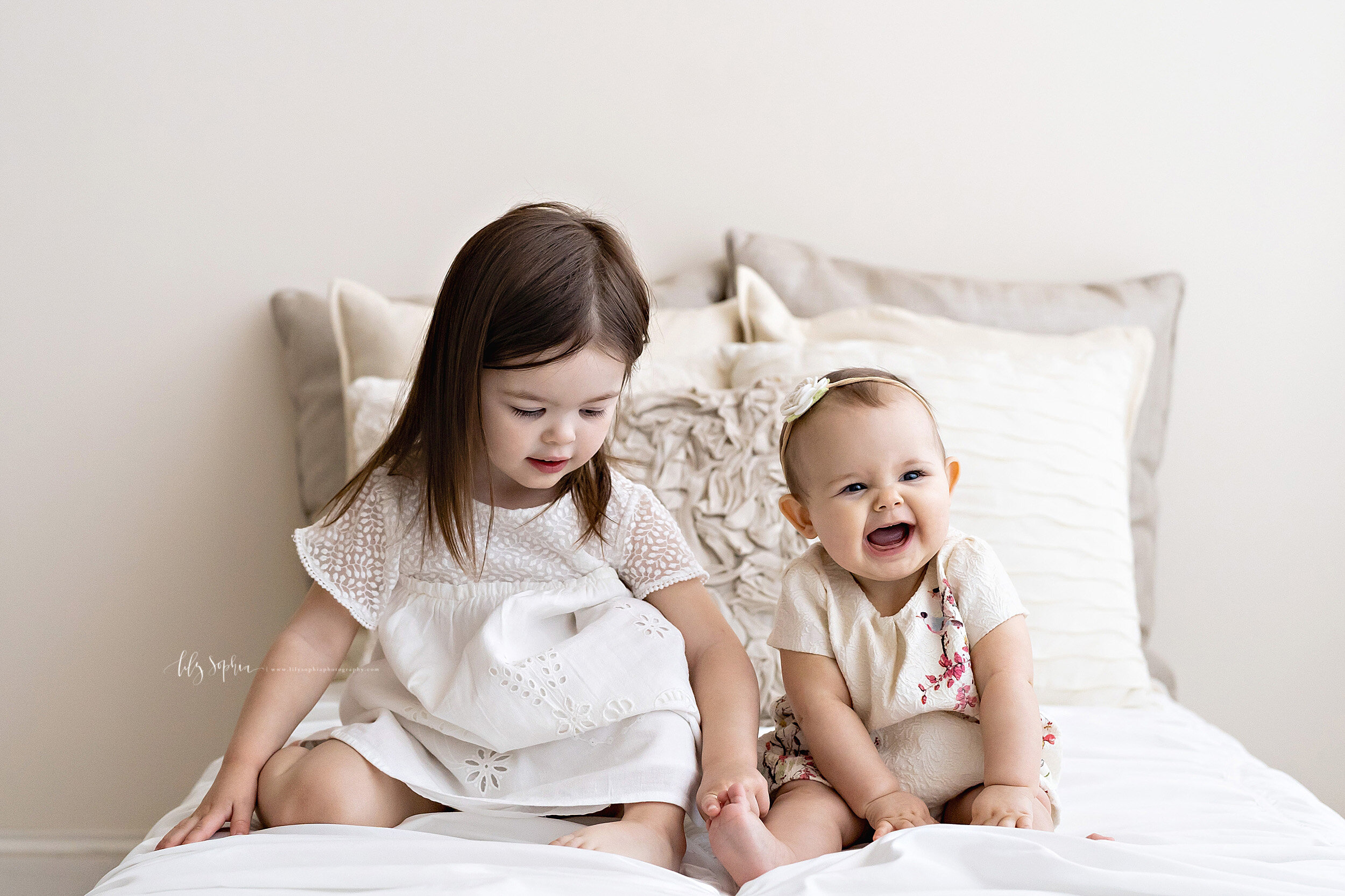  Family photo of sibling girls sitting next to each other on a bed in front of a natural light window with the baby wearing a rose headband and laughing because her sister is tickling her toes taken in a Midtown studio in Atlanta. 