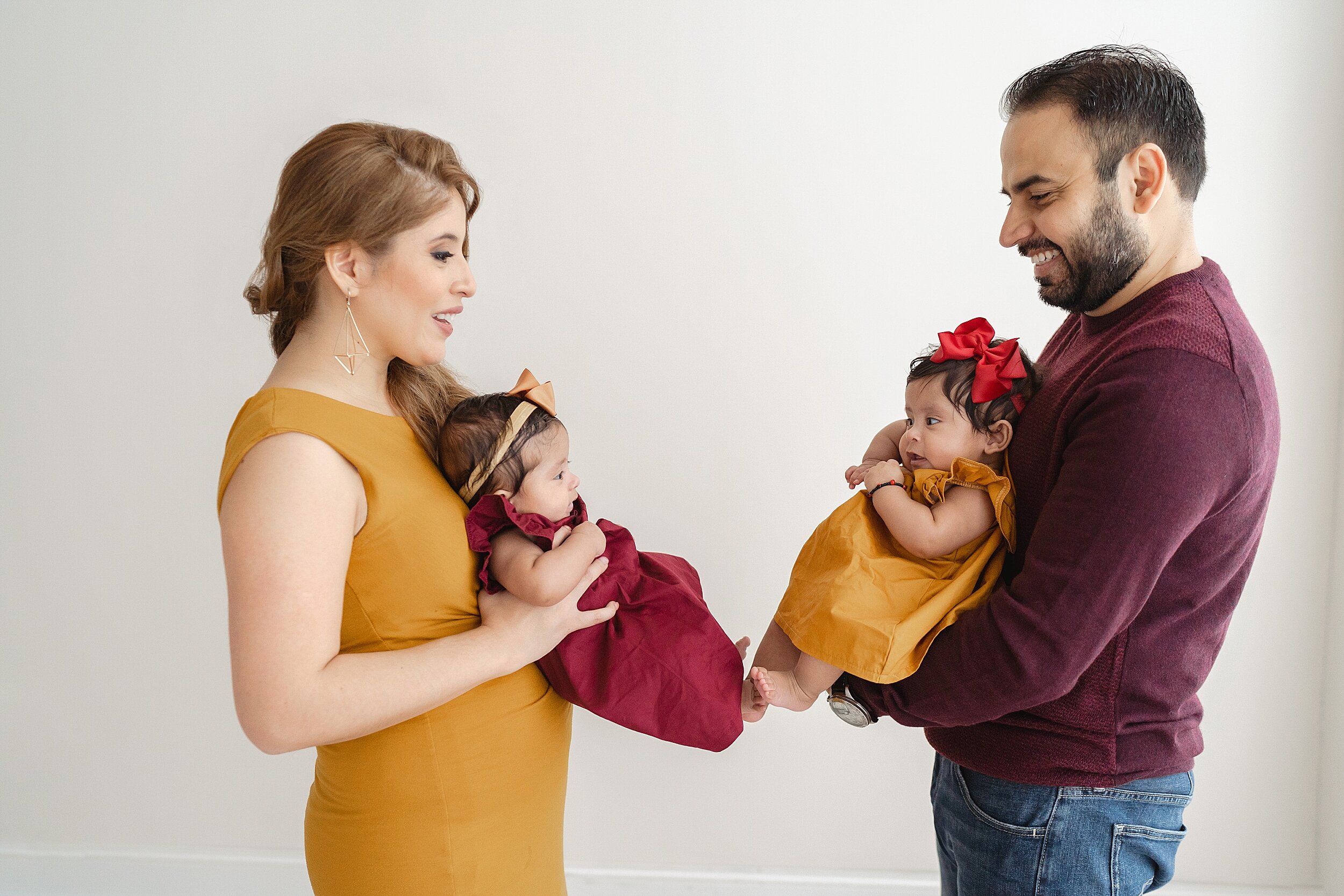  Newborn photograph of a mother holding one of her twin baby girls in front of her and facing her husband while the father holds the other twin as the twins interact with one another and touch toes taken in a natural light studio in the Brookhaven ar
