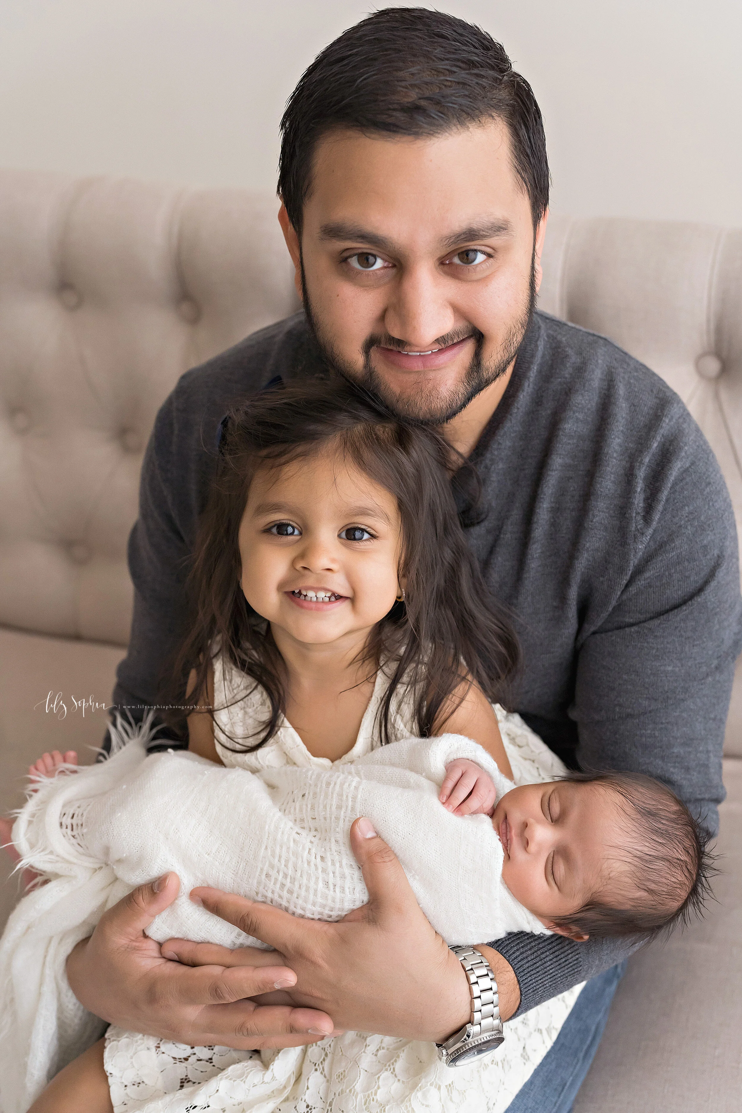 Newborn photograph of an Indian dad holding his toddler daughter on his lap while she holds her newborn baby brother on her lap as they sit on a sofa in natural light in a studio in the Ansley Park area of Atlanta, Georgia.