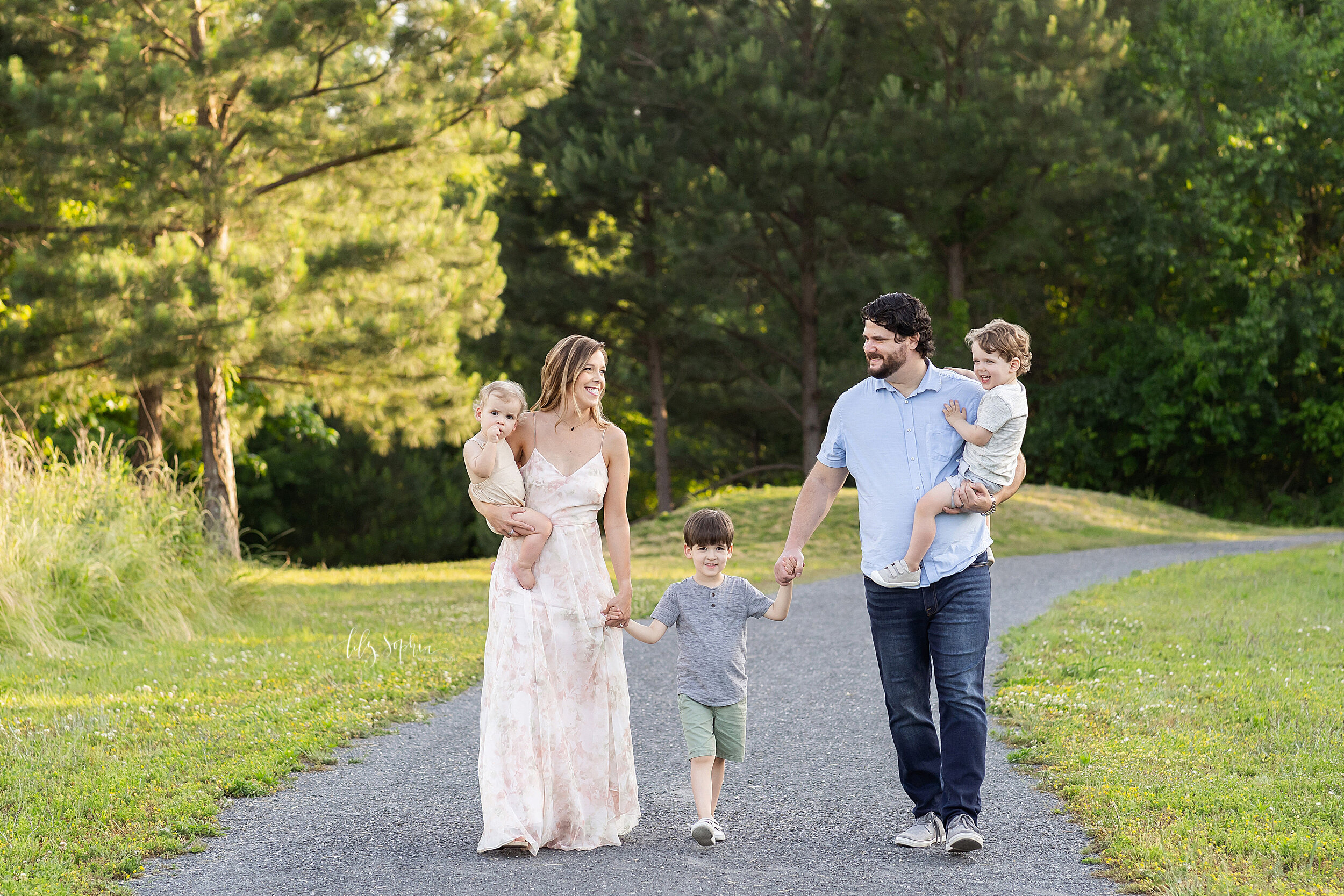  Family photo of a mom holding her baby boy on her right hip and dad holding his toddler son on his left hip with both parents holding the hands of their eldest son during the summer as the family walks along a path in an Atlanta park at sunset. 