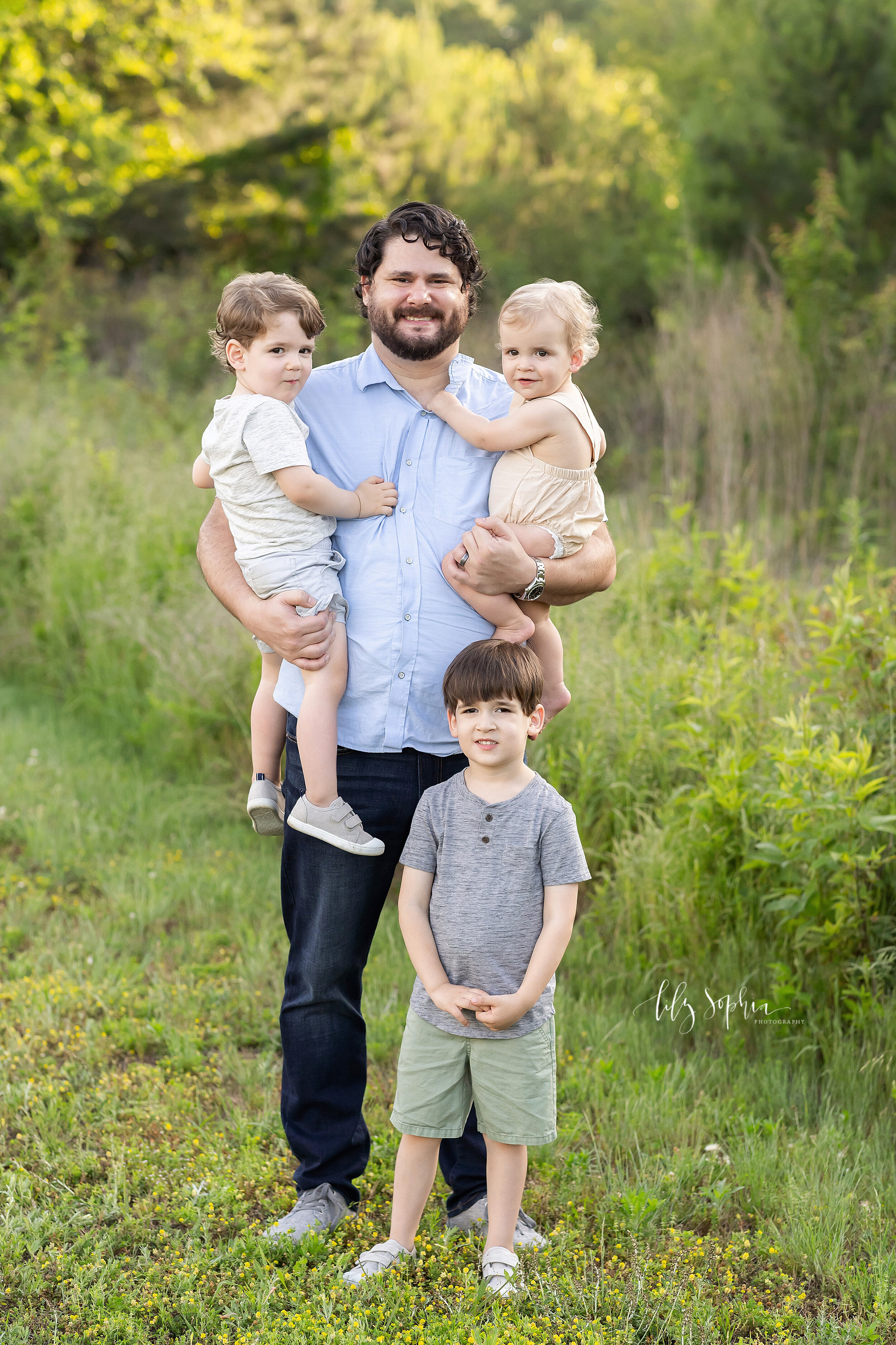  Family photo of a father holding his baby son in his right arm and his toddler son in his left arm while he stands with his eldest son in a field in Atlanta at sunset in the summer. 