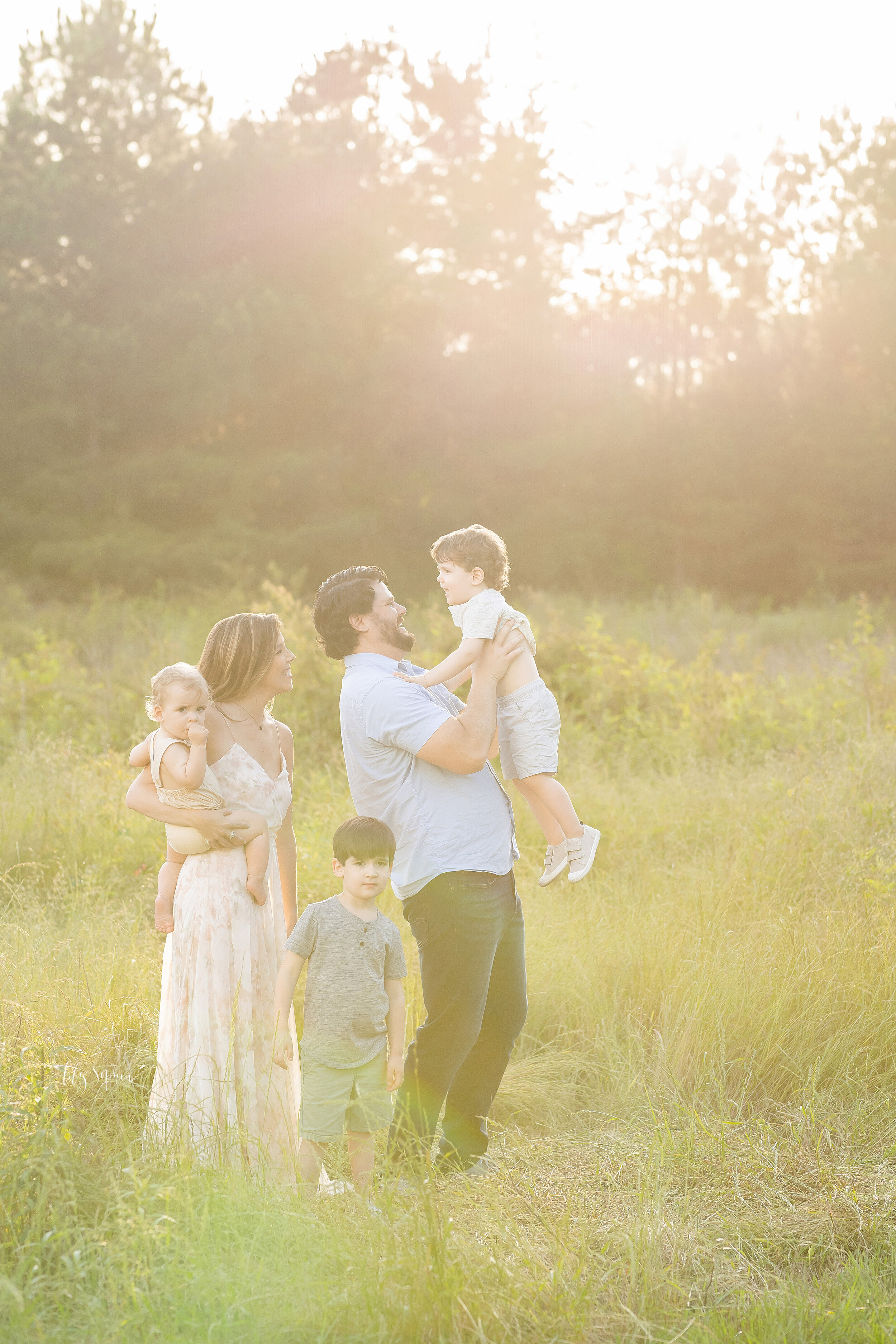  Family photo of five with mom holding her baby son on her hip, dad lifting his toddler son above his head and the couple’s eldest son quietly standing between them taken at sunset in natural light in a field near Atlanta, Georgia. 