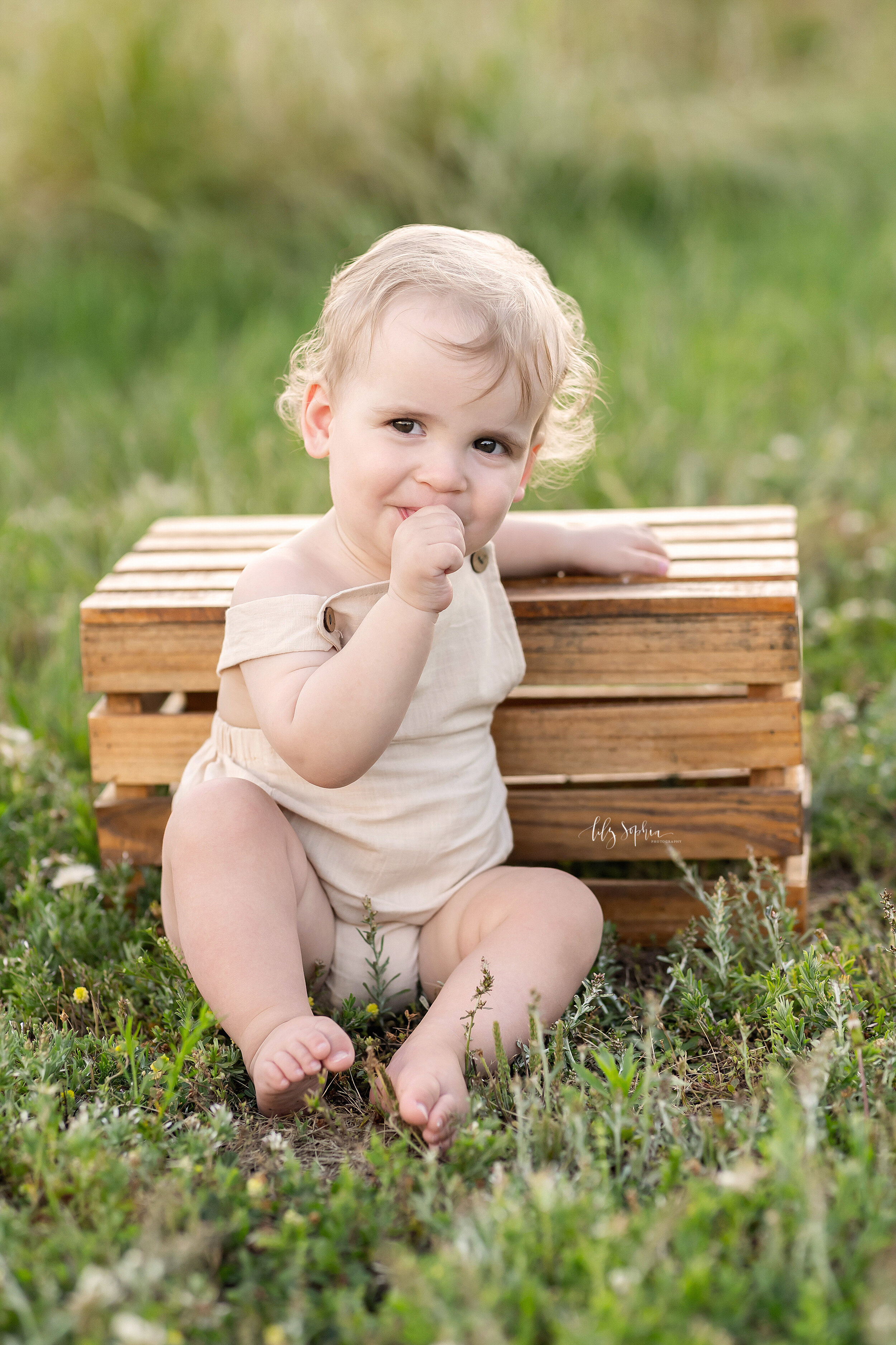  Family photo of a toddler boy as he sits leaning on a wooden box with his right thumb in his mouth during the summer in a field near Atlanta, Georgia at sunset. 
