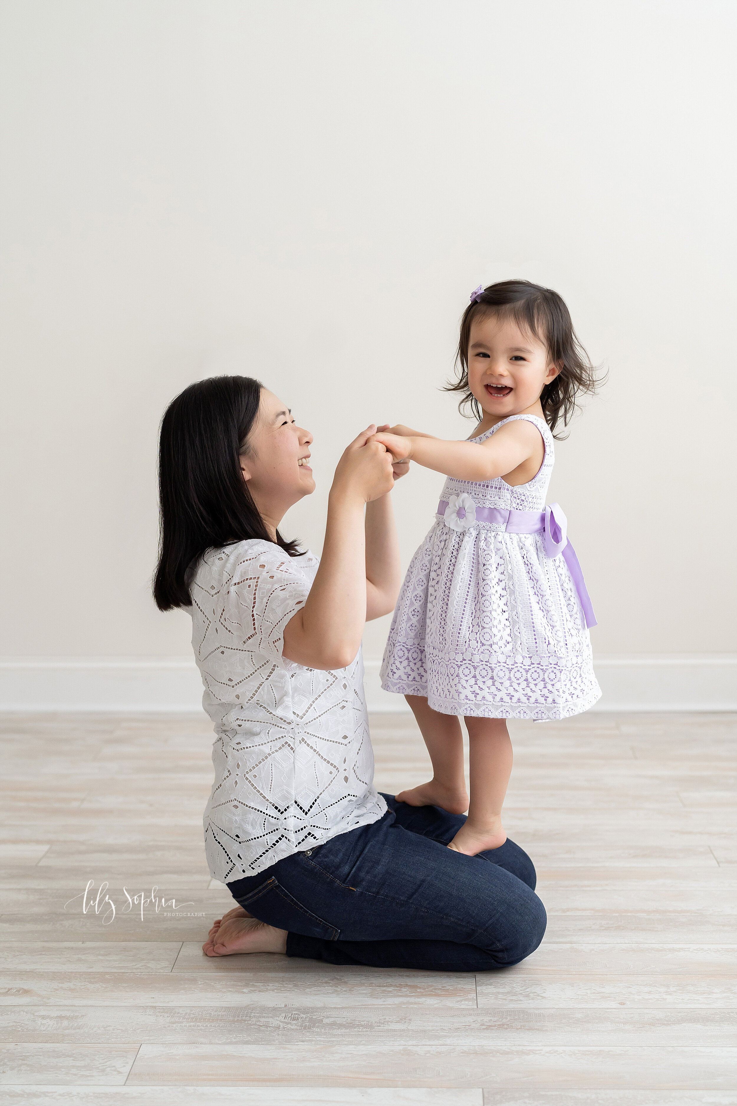  Family photo of an Asian mother sitting on the floor of a natural light studio in a white short-sleeved shirt and blue jeans with her two-year old daughter dressed in a sleeveless lilac dress covered in lace standing on her knees and holding her han