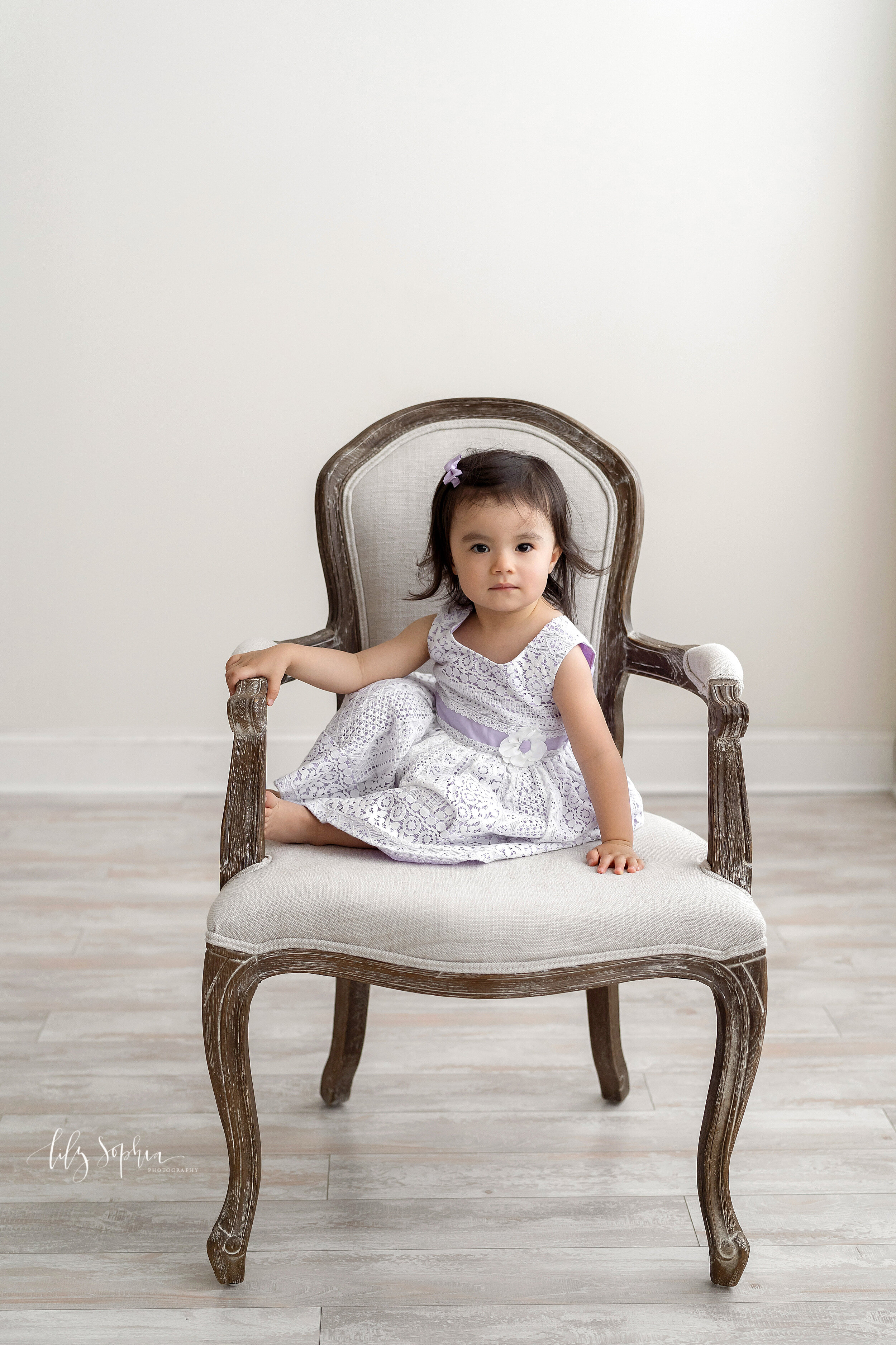  Milestone photo of an Asian two-year old girl as she sits in an armchair in a lilac and lace dress in a studio near the Buckhead area of Atlanta in natural light. 