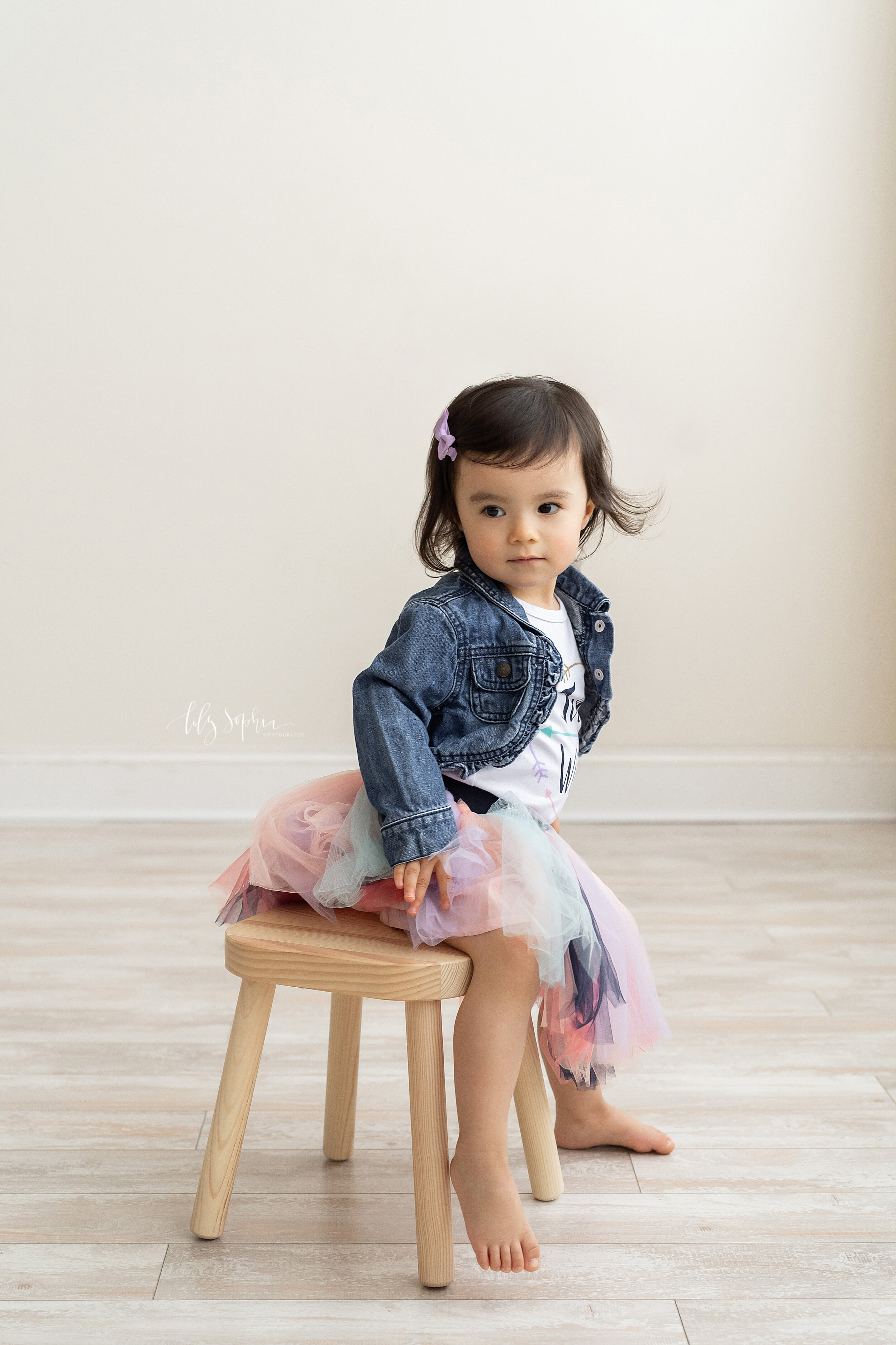  Milestone photo of an Asian two-year old as she sits on a wooden stool wearing a multi-colored tuille ballerina skirt a t-shirt and a jean jacket with a lilac bow in her hair taken in a studio in the Candler Park area of Atlanta, Georgia in natural 